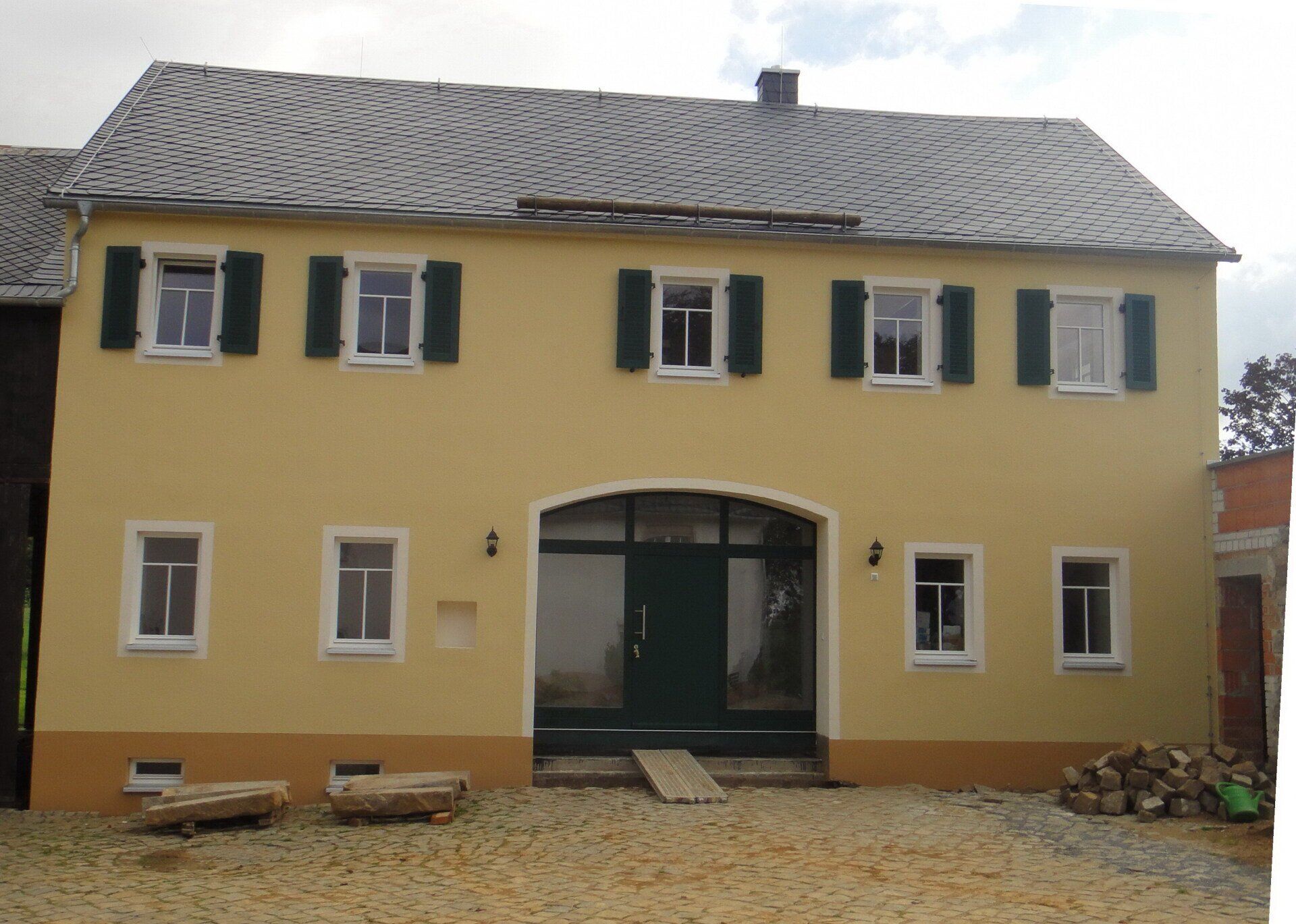 Gelbes zweistöckiges Gebäude mit grünen Fensterläden, gewölbtem Eingang und grauem Dach. Kieshof mit Brennholz.