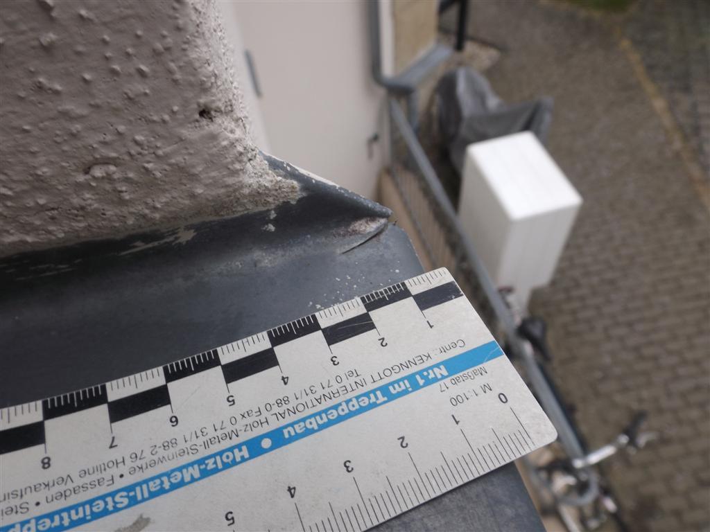 Nahaufnahme einer dunklen Metallkante neben einer Wand; ein Lineal wird über das Metall gelegt, um es zu messen.