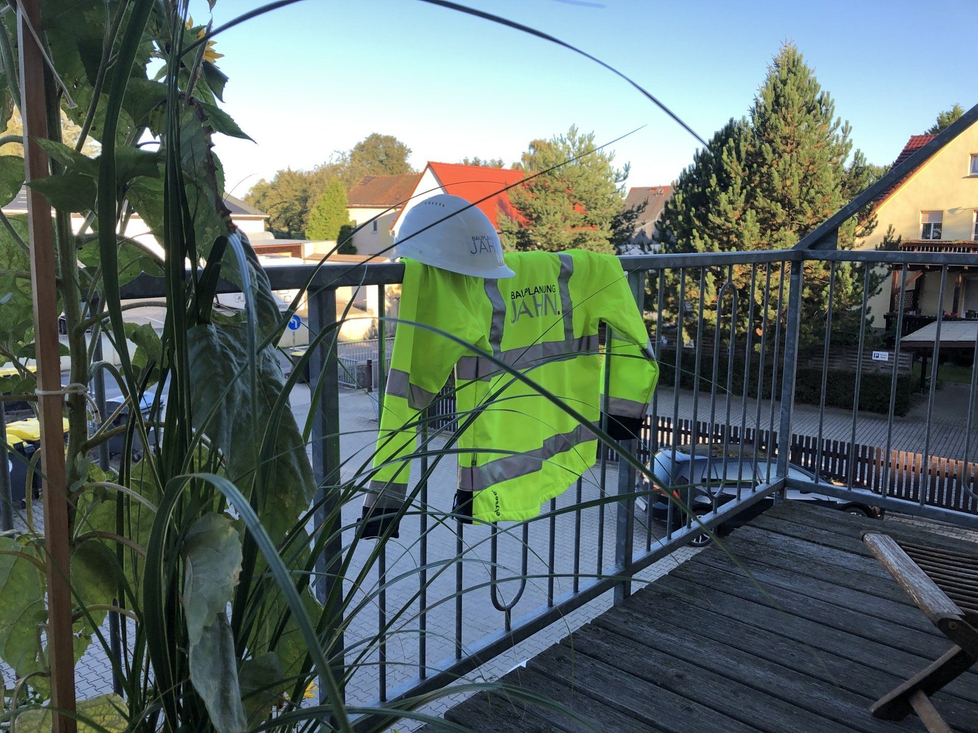 Eine Warnweste und ein weißer Schutzhelm hängen an einem Balkongeländer; im Hintergrund sind Wohnhäuser zu sehen.