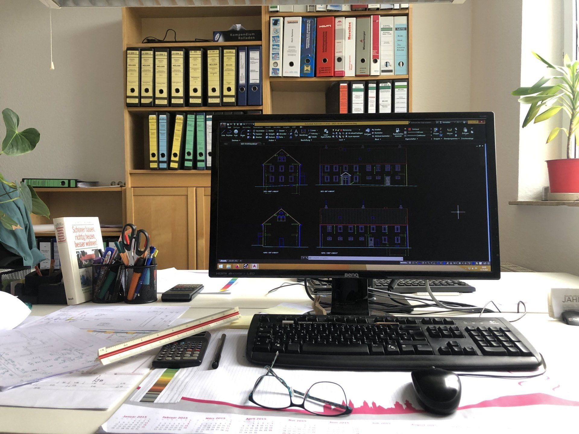 Ein Architekturbüro mit einem Computer, auf dem Baupläne und Dateien angezeigt werden, und einem Regal mit Ordnern.