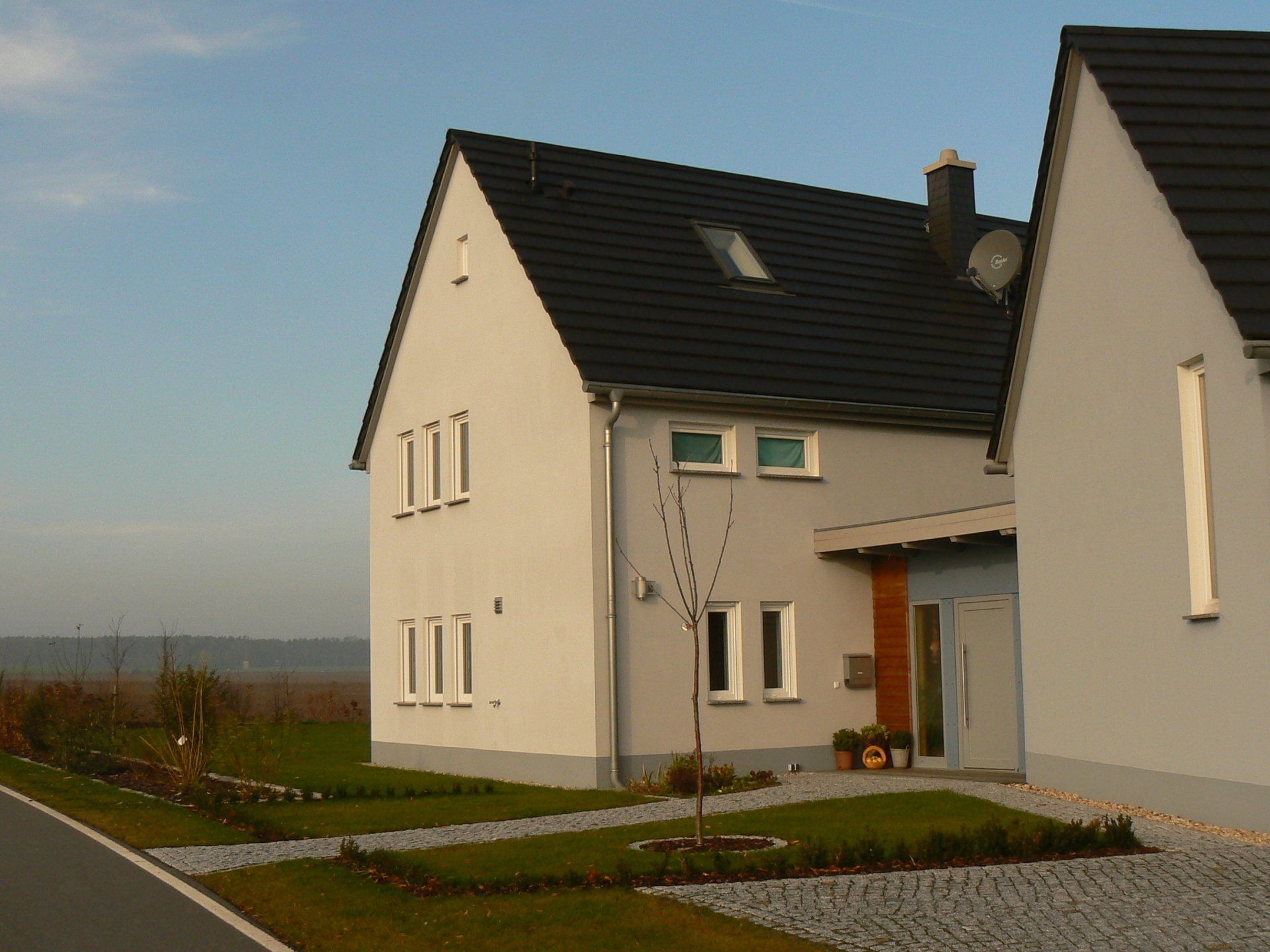 Zweistöckiges graues Haus mit schwarzem Dach, ein kleiner Hof mit gepflasterter Auffahrt und im Hintergrund klarer Himmel.