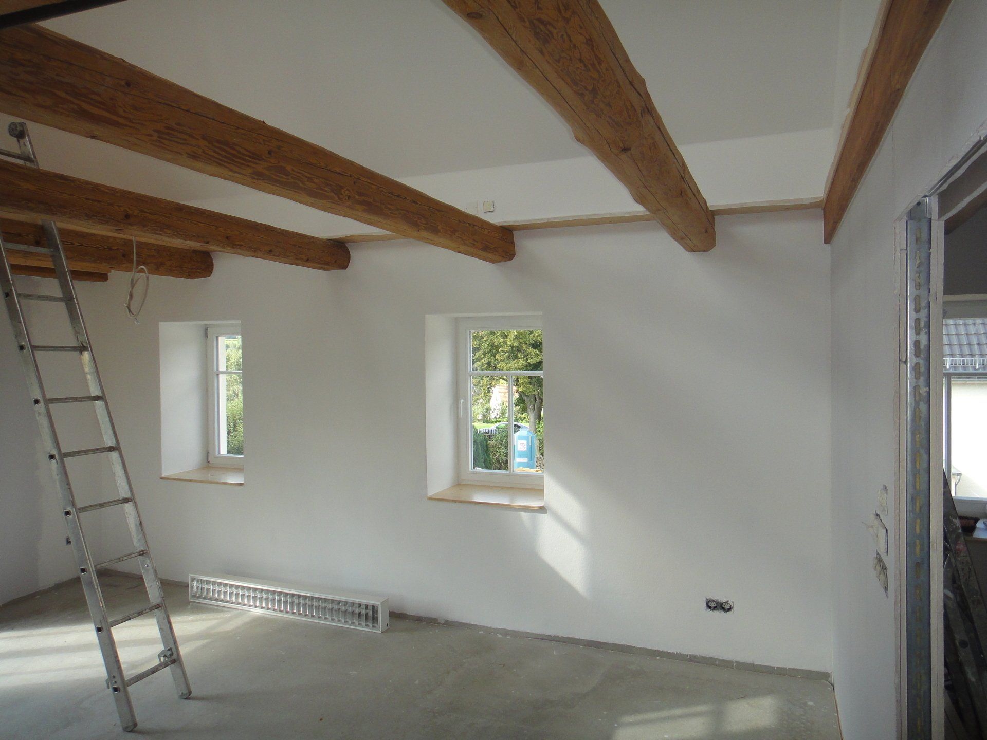 Innenraum mit freiliegenden Holzbalken, zwei Fenstern, Leiter und unverputzten Wänden.
