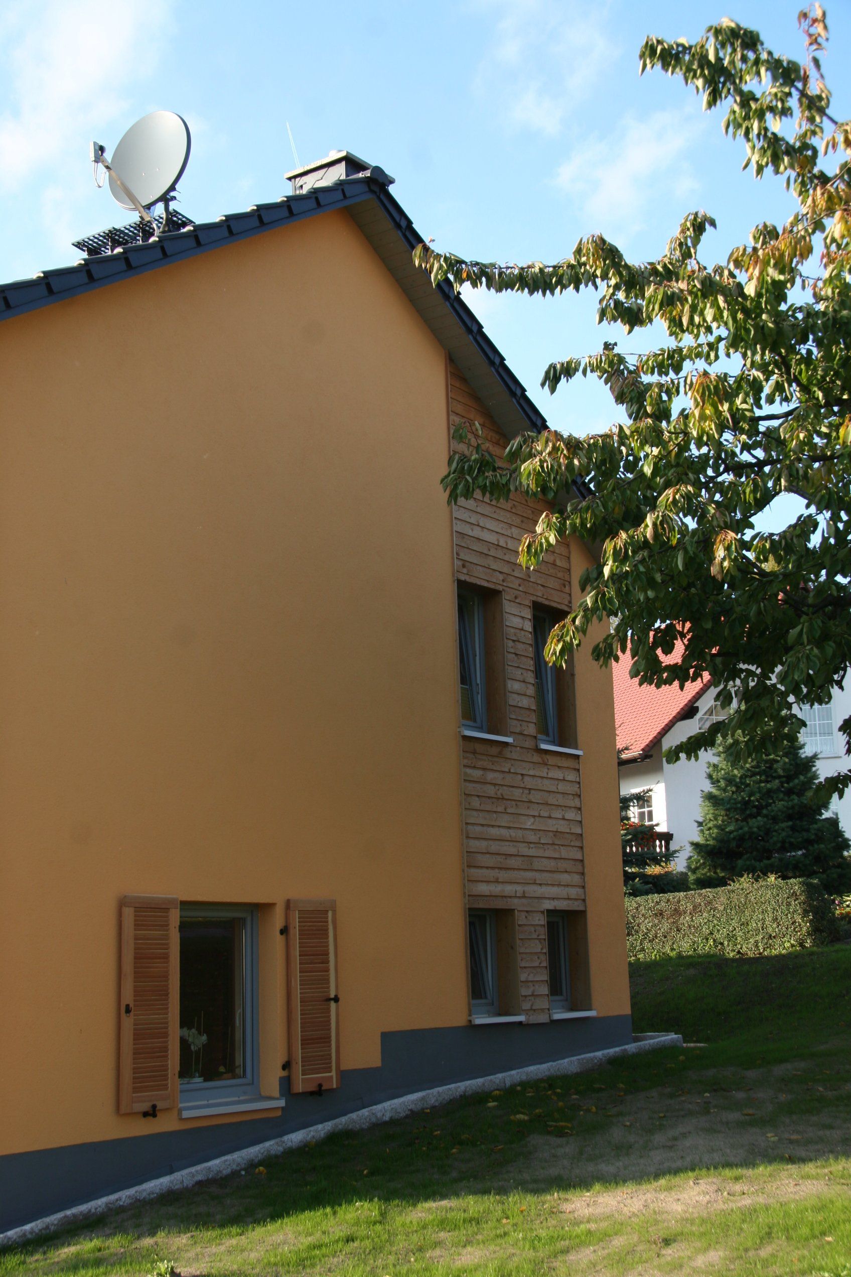 Seite eines zweistöckigen Gebäudes mit orangefarbenem Stuck und Ziegeln, Satellitenschüssel auf dem Dach, Fensterläden und einem Rasenhof.