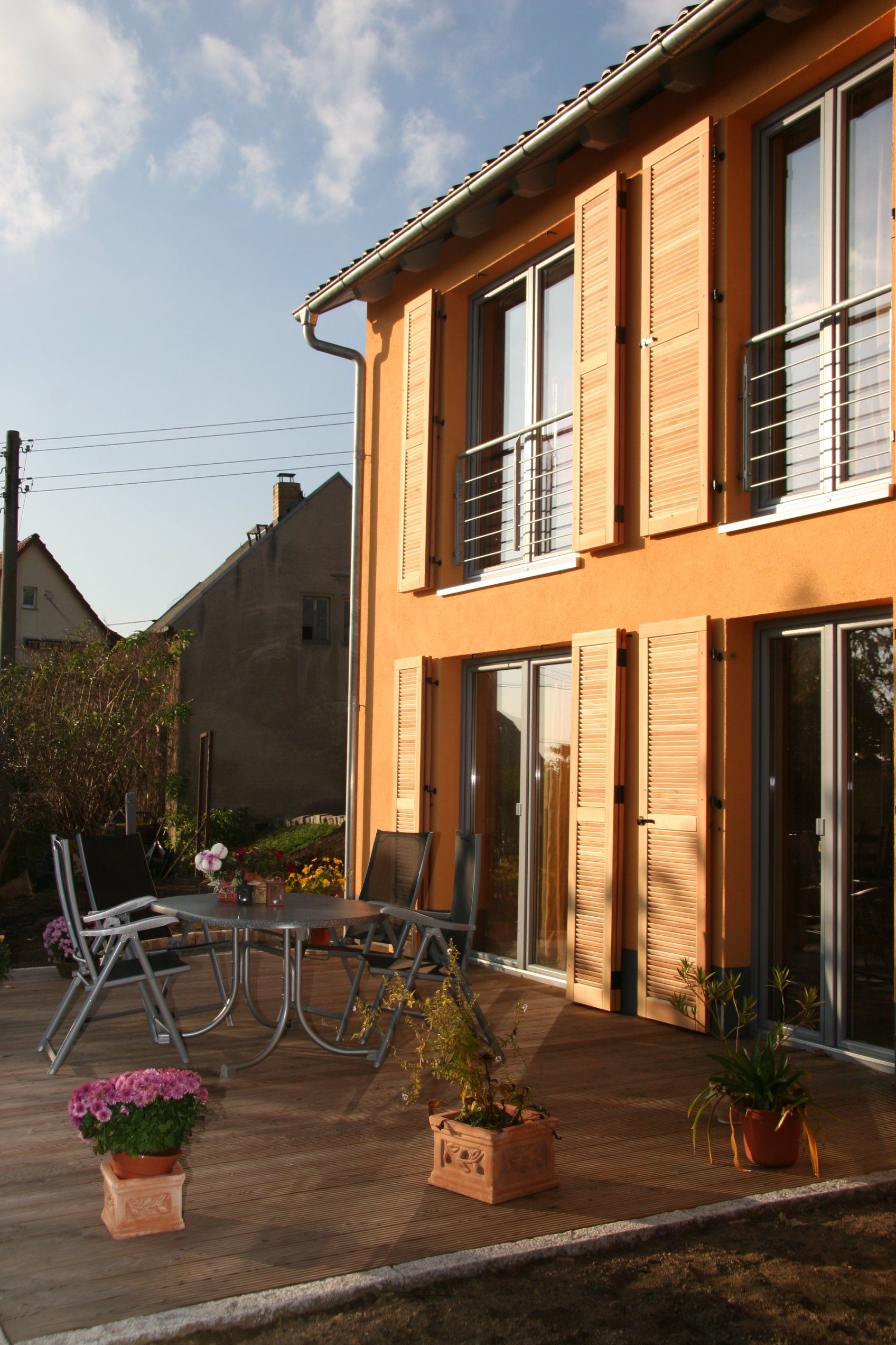 Orangefarbenes Haus mit Holzfensterläden, Terrasse mit Tisch, Stühlen und Topfpflanzen.