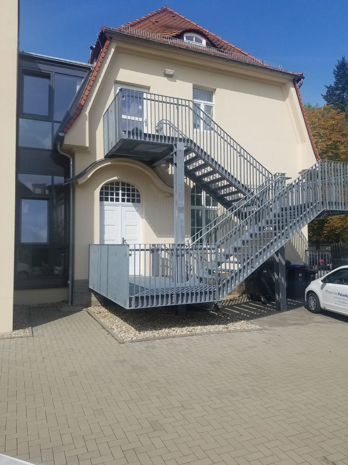 Außenansicht eines zweistöckigen Gebäudes mit einer metallenen Feuertreppe. Ein weißes Auto ist in der Nähe geparkt.
