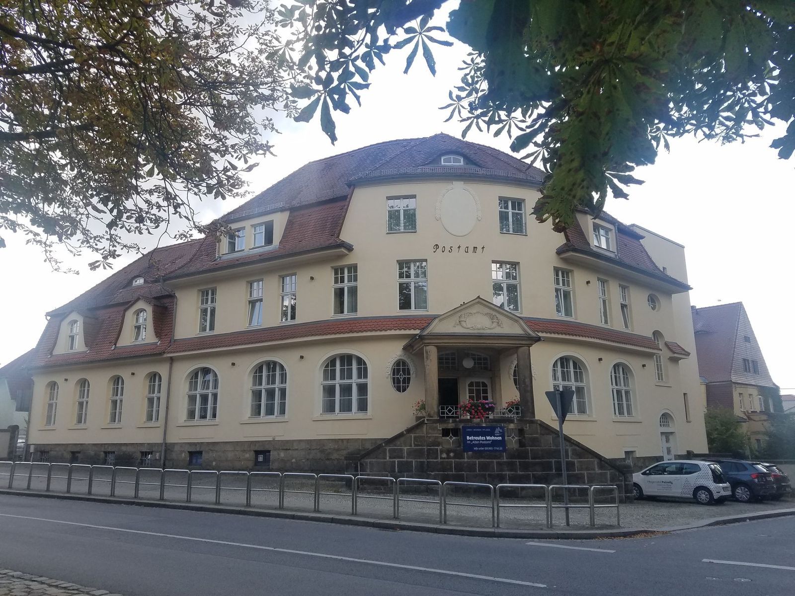 Beiges Gebäude mit rotem Ziegeldach und mehreren Fenstern, Straße davor, Autos rechts geparkt.