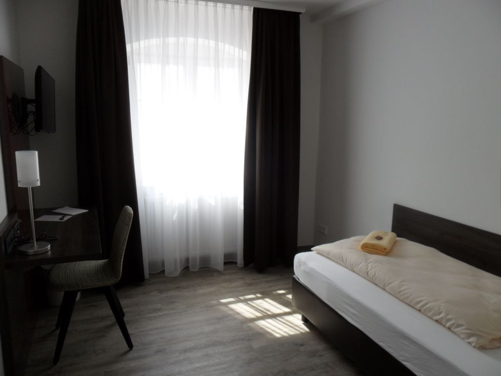 Hotelzimmer mit Bett, Schreibtisch, Stuhl, Fenster, Vorhängen und Fernseher.