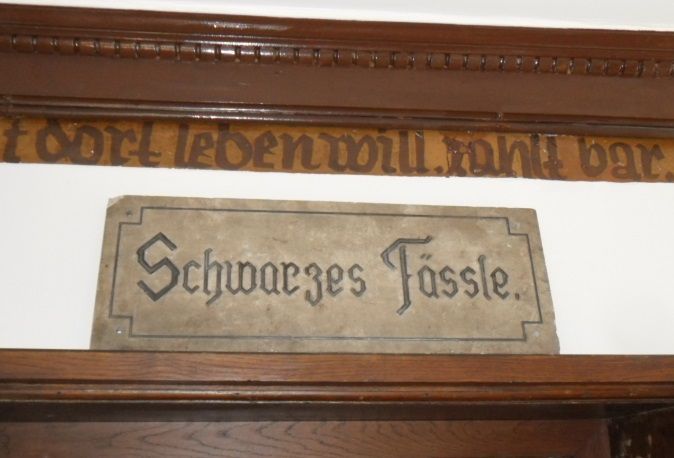 Über einem Holztürrahmen mit deutscher Beschriftung prangt ein Schild mit der Aufschrift „Schwarzes Fässle“.