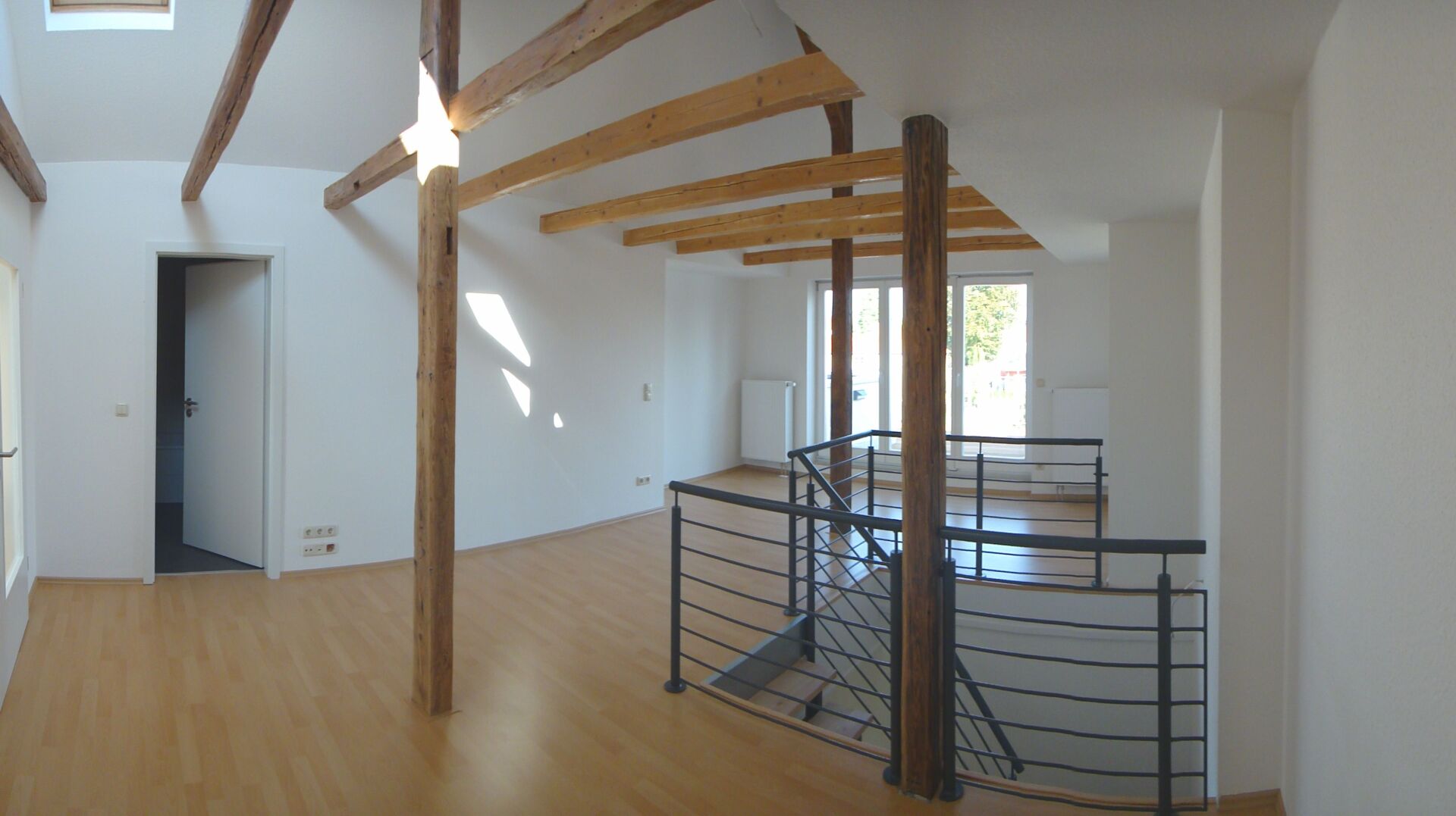 Innenansicht eines Zimmers mit Holzbalken und Treppe. Helle Beleuchtung, weiße Wände und Holzboden.