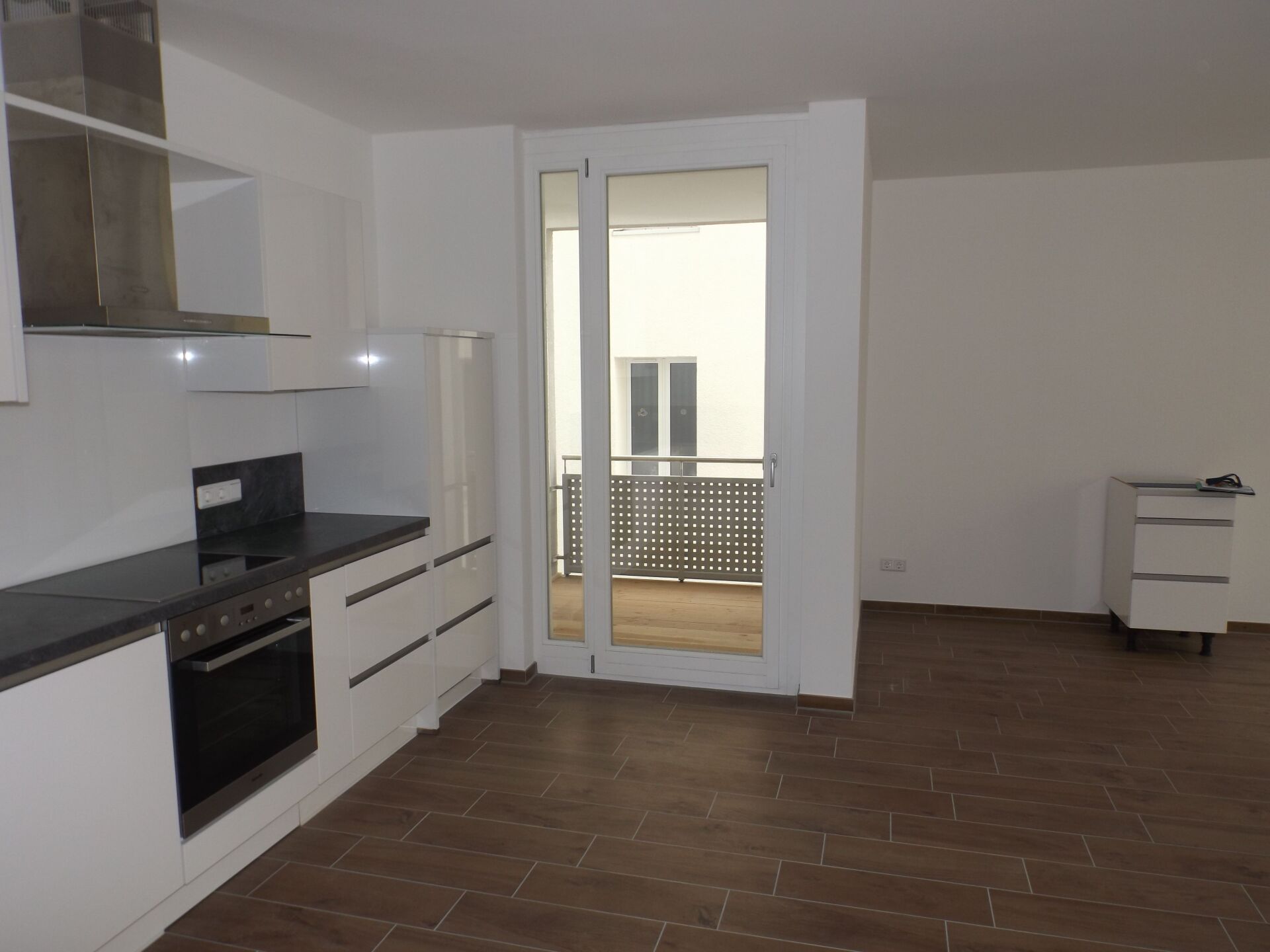 Moderne Küche mit weißen Schränken, dunklen Arbeitsplatten und Balkontür. Brauner Fliesenboden.