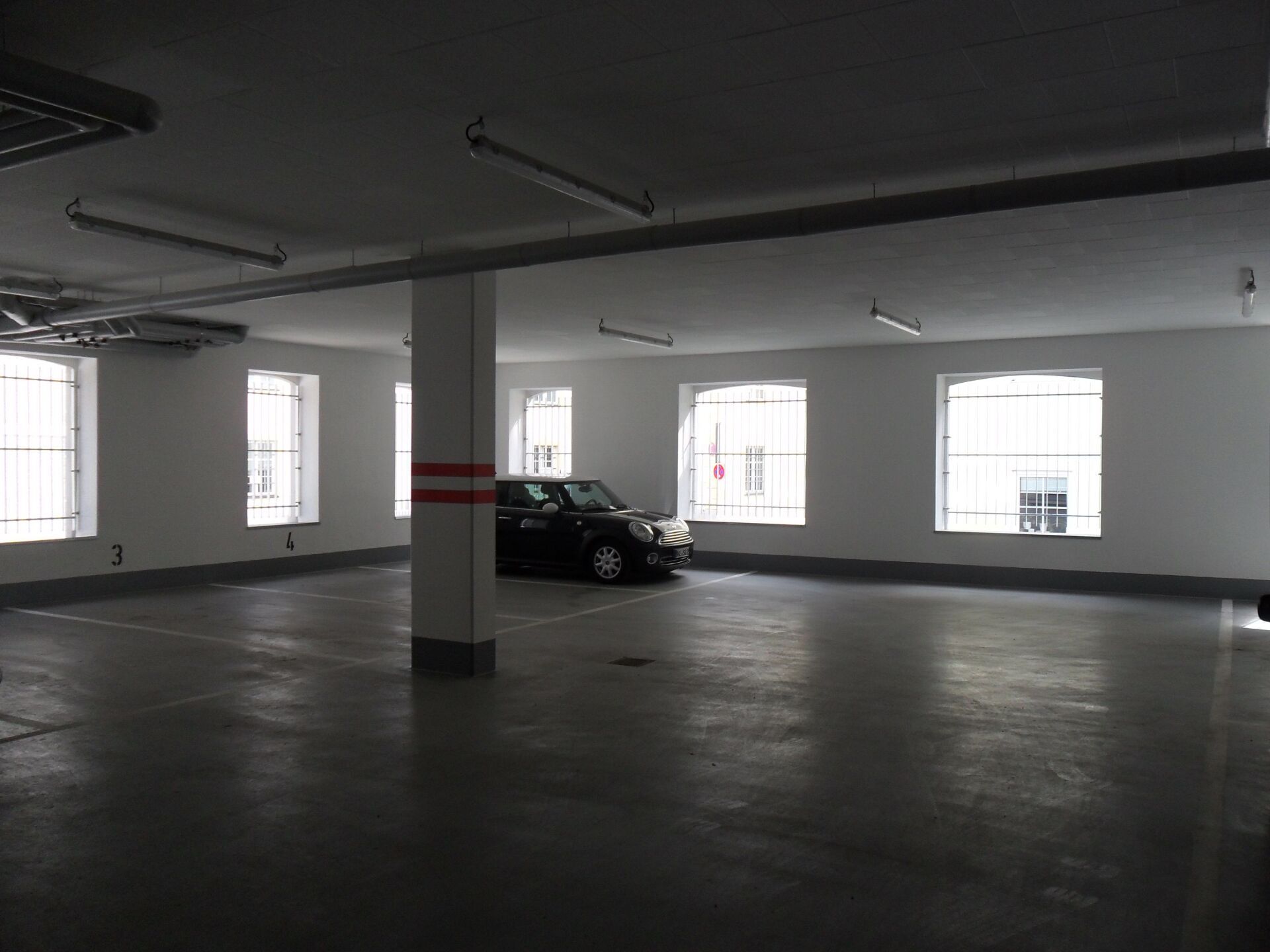 Leeres, graues Parkhaus mit einem schwarzen Auto, das in der Nähe einer Säule und der Fenster geparkt ist.