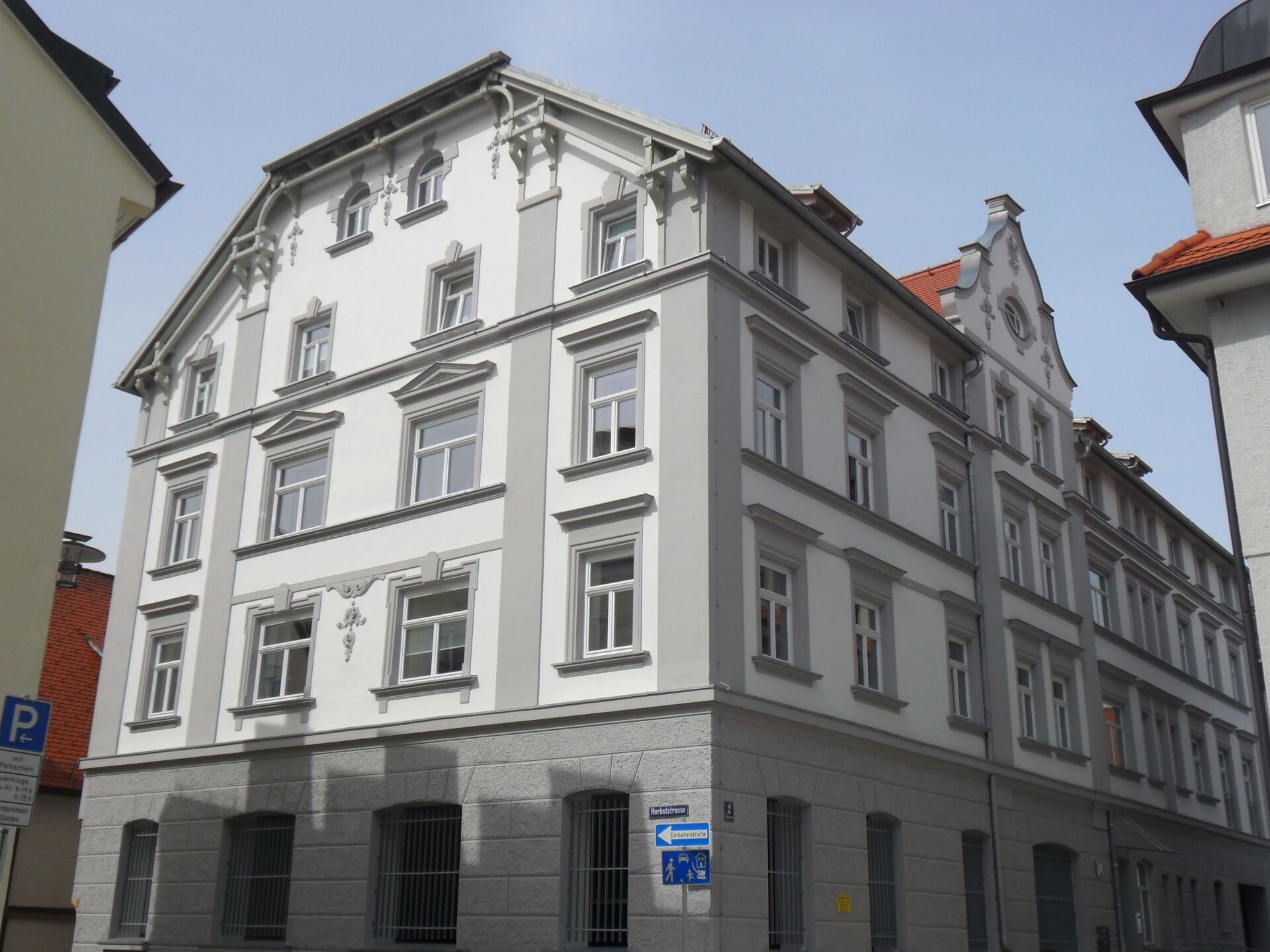 Vierstöckiges Gebäude mit grau-weißer Fassade, zahlreichen Fenstern und einem roten Ziegeldach unter blauem Himmel.