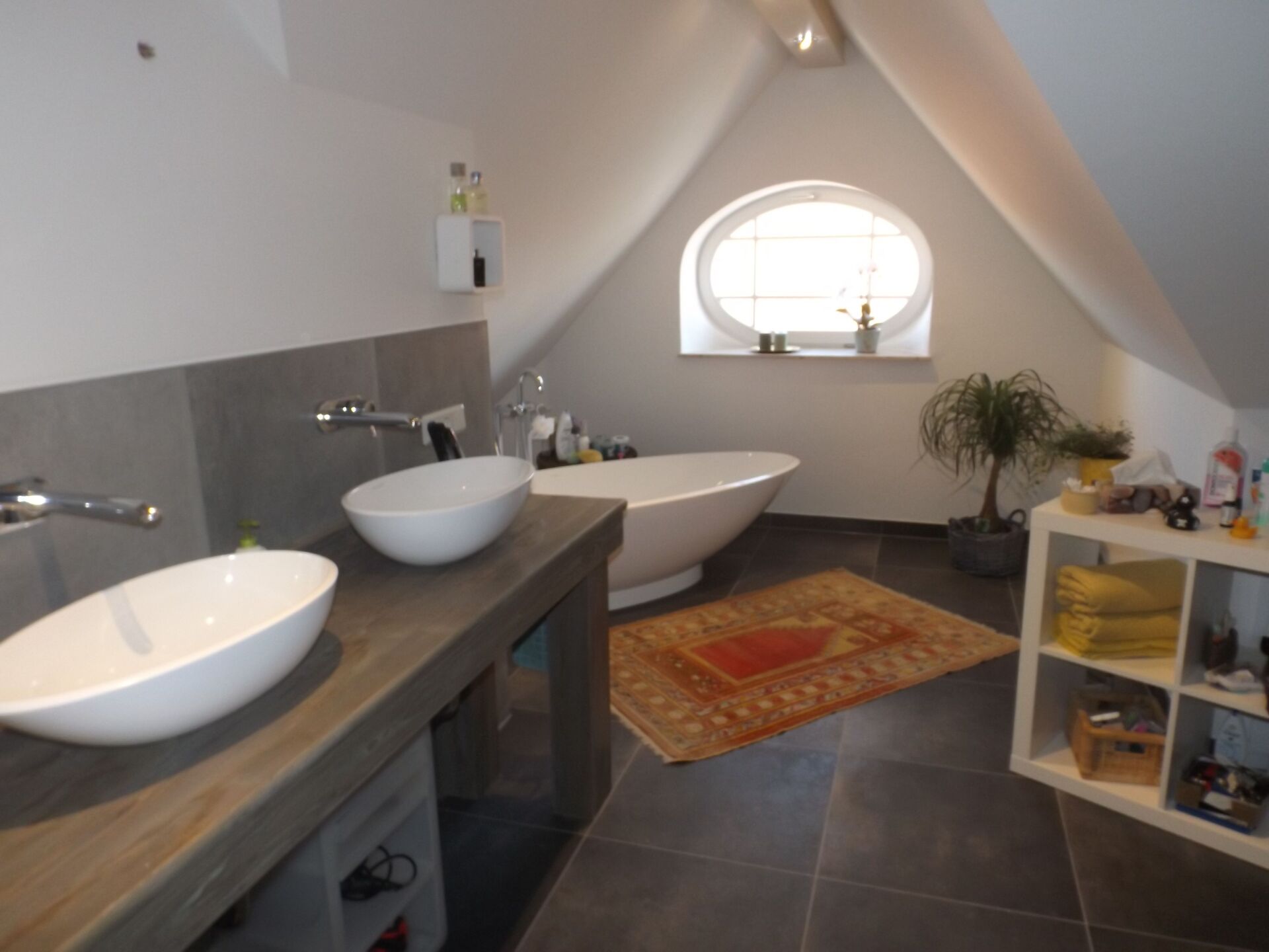 Badezimmer mit zwei Waschbecken, einer freistehenden Badewanne und einem kleinen Teppich unter einem Fenster in einem Raum mit Dachschräge.