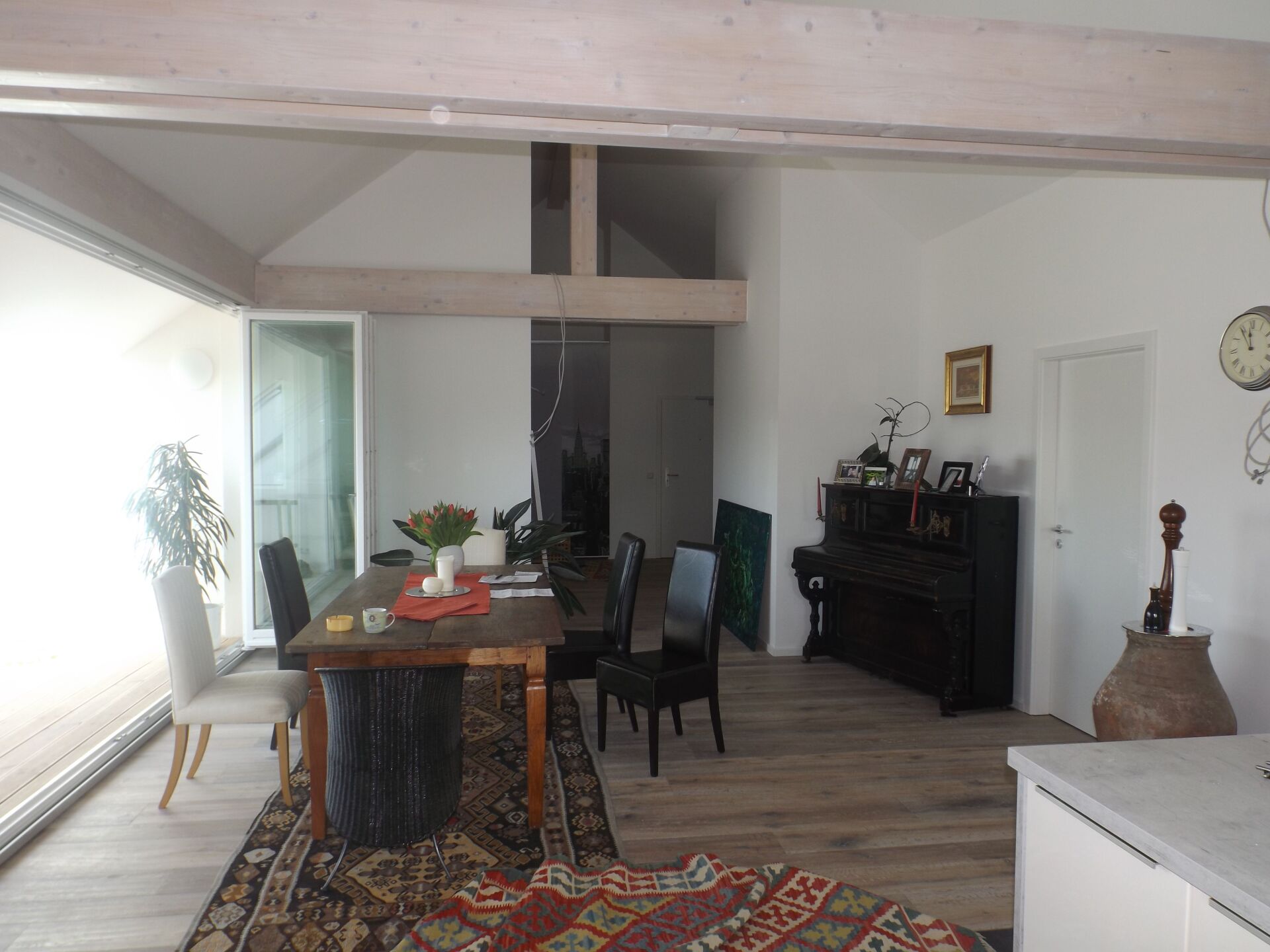 Essbereich mit Holztisch, Teppich und Klavier, dazu weiße Wände und Holzbalken.
