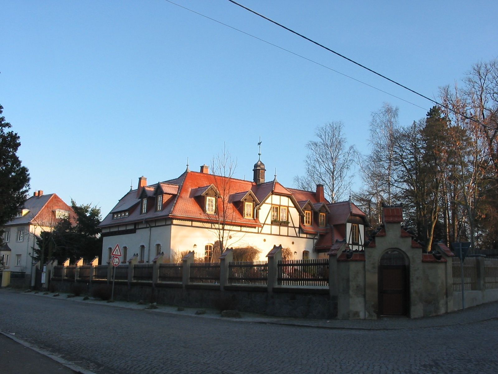 Ein mehrstöckiges Gebäude mit rotem Ziegeldach und Steinmauer, gelegen an einer Straße mit klarem Himmel.