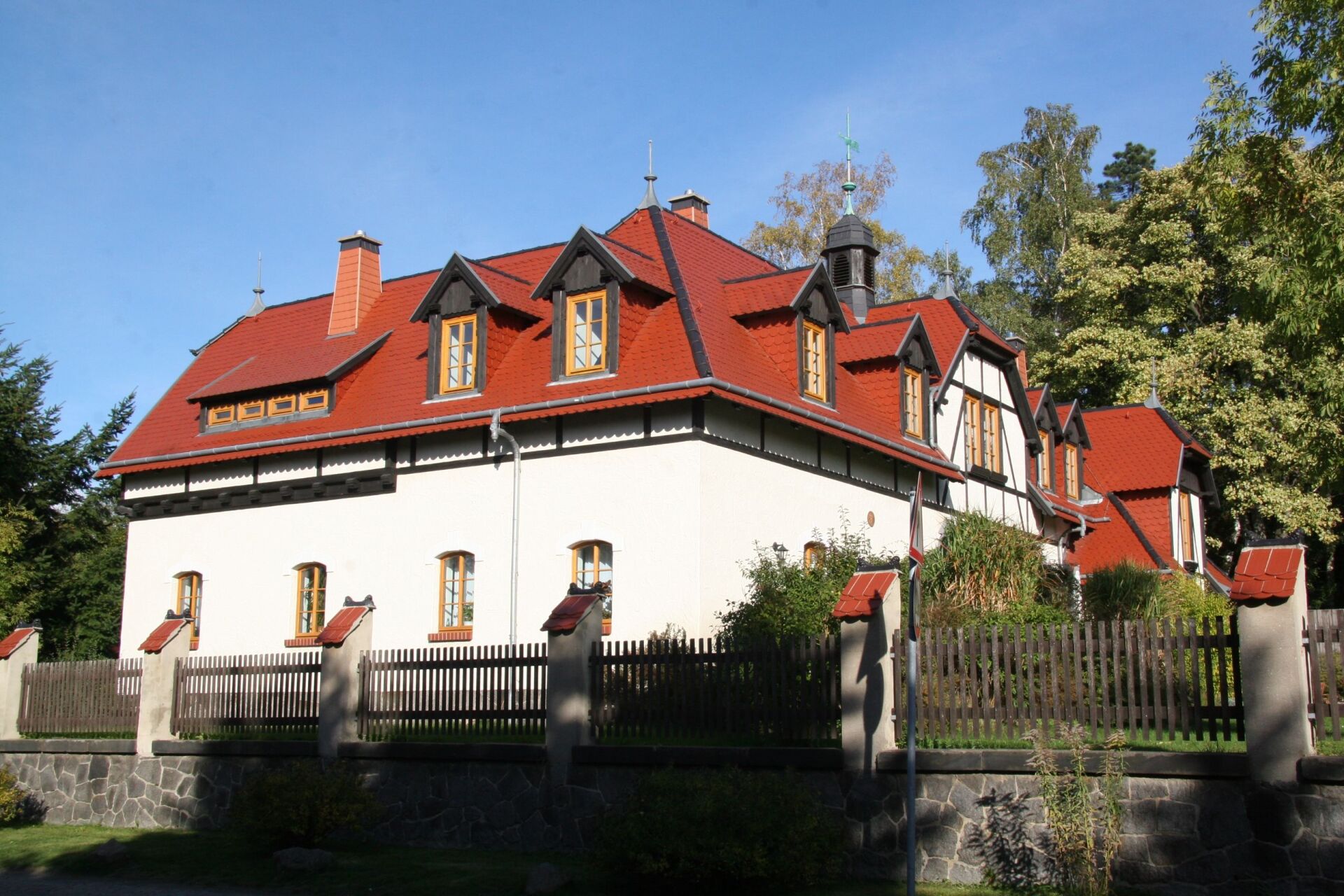 Zweistöckiges Haus mit rotem Dach, weißen Wänden und Holzverzierungen, hinter einem Holzzaun gelegen.