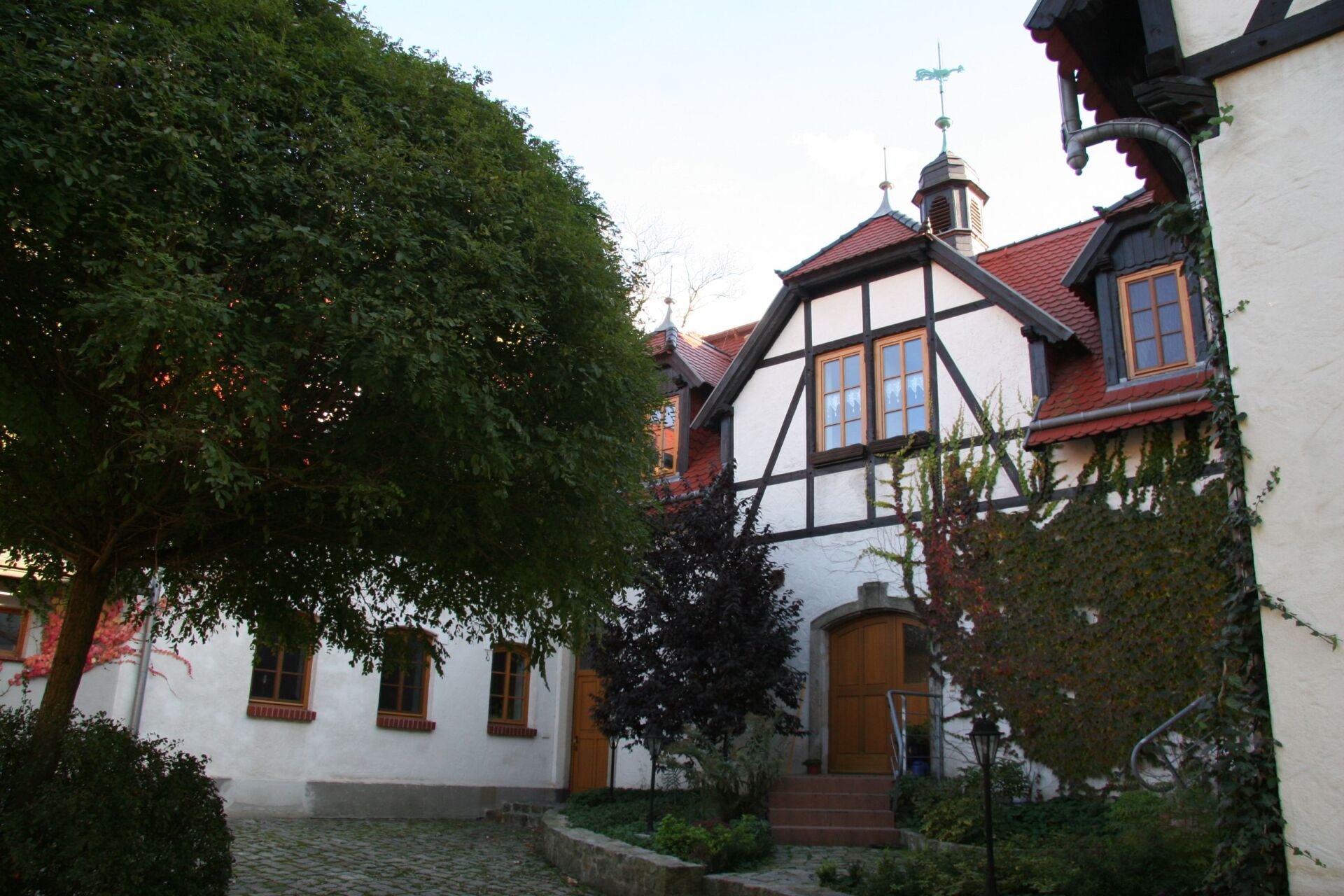 Fachwerkhaus mit rotem Dach, braunen Türen und grünem Efeu. Innenhof mit großem Baum.