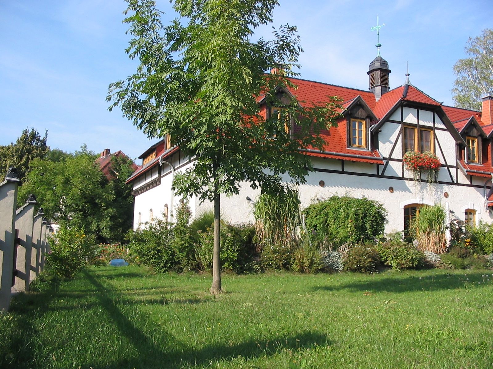Ein weißes Fachwerkhaus mit rotem Ziegeldach; davor grünes Gras und Bäume an einem sonnigen Tag.