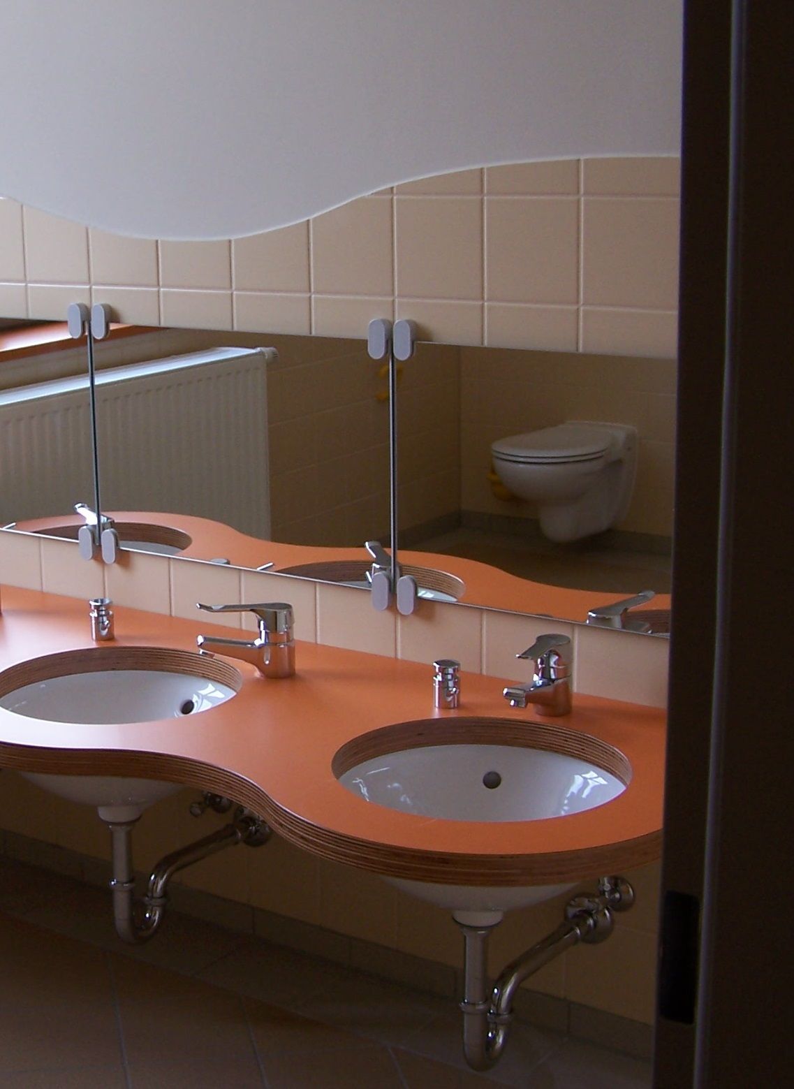 Öffentliche Toilette mit zwei runden Waschbecken, geschwungener Arbeitsplatte und einer Toilette im Hintergrund. Beige- und Orangetöne.
