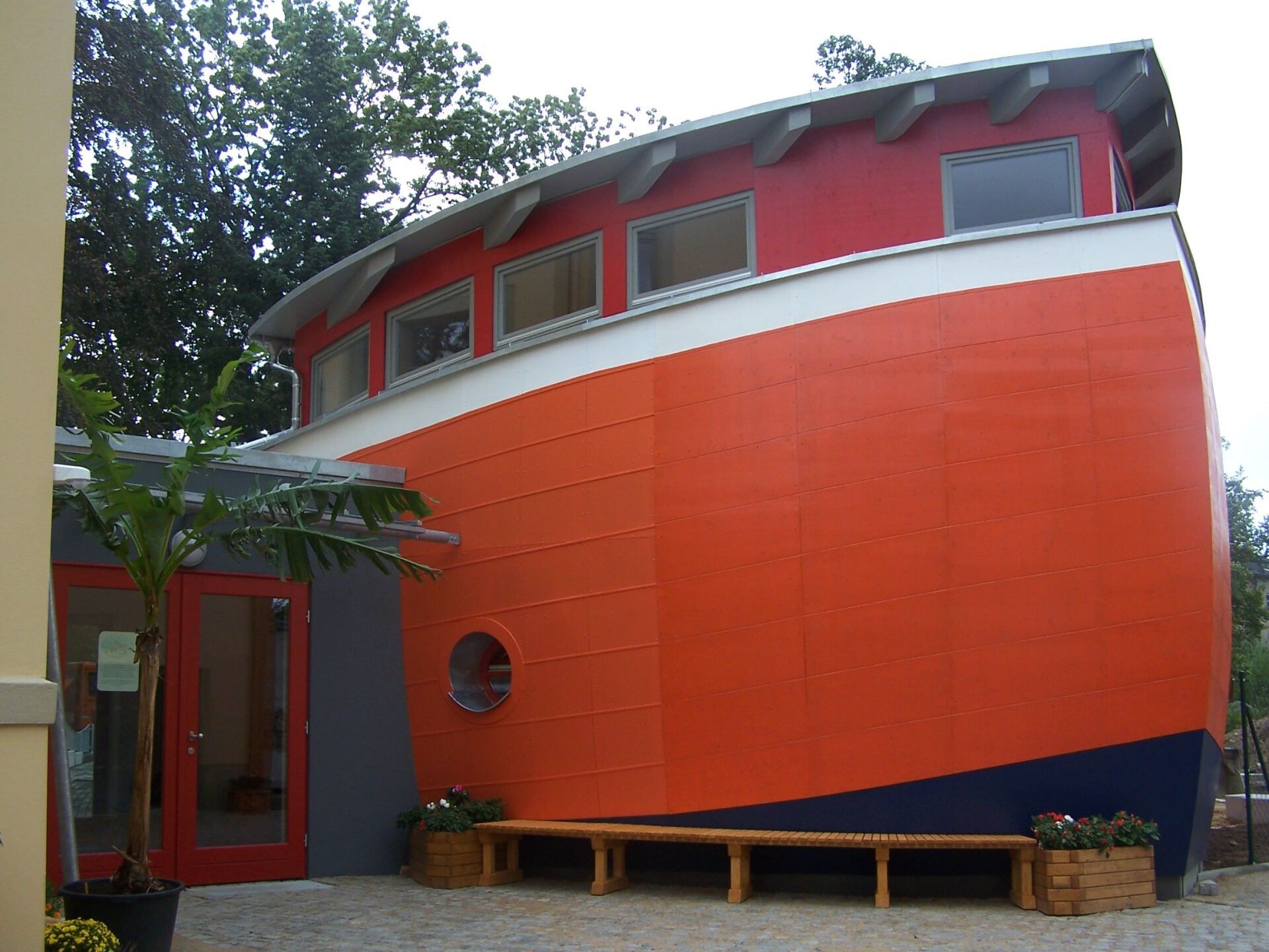 Orange-rotes Gebäude mit bootsähnlichem Design, rundem Fenster, Bank und roter Tür.