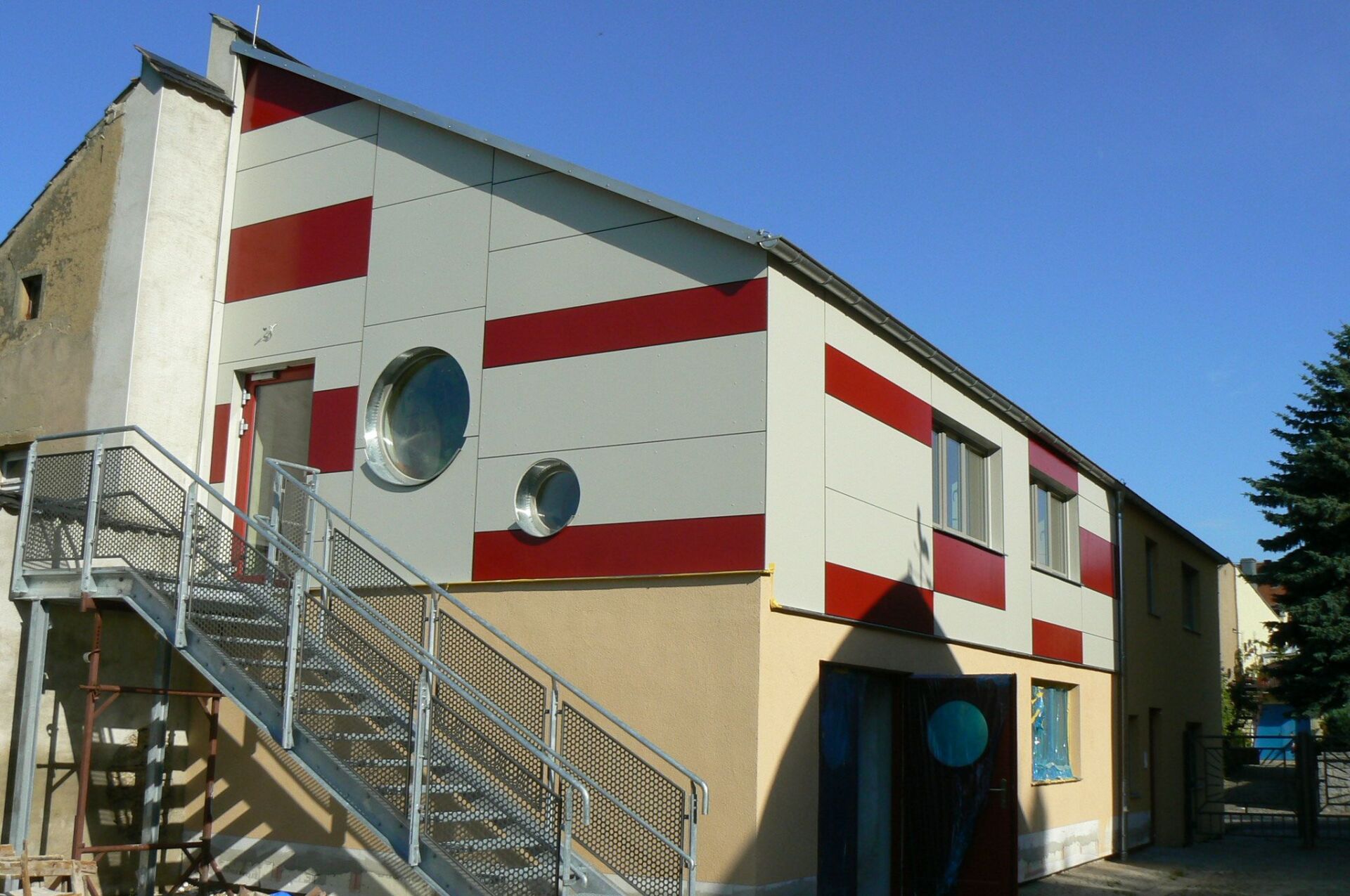 Außenansicht eines Gebäudes mit roten und weißen Paneelen. Eine Metalltreppe führt ins Obergeschoss.