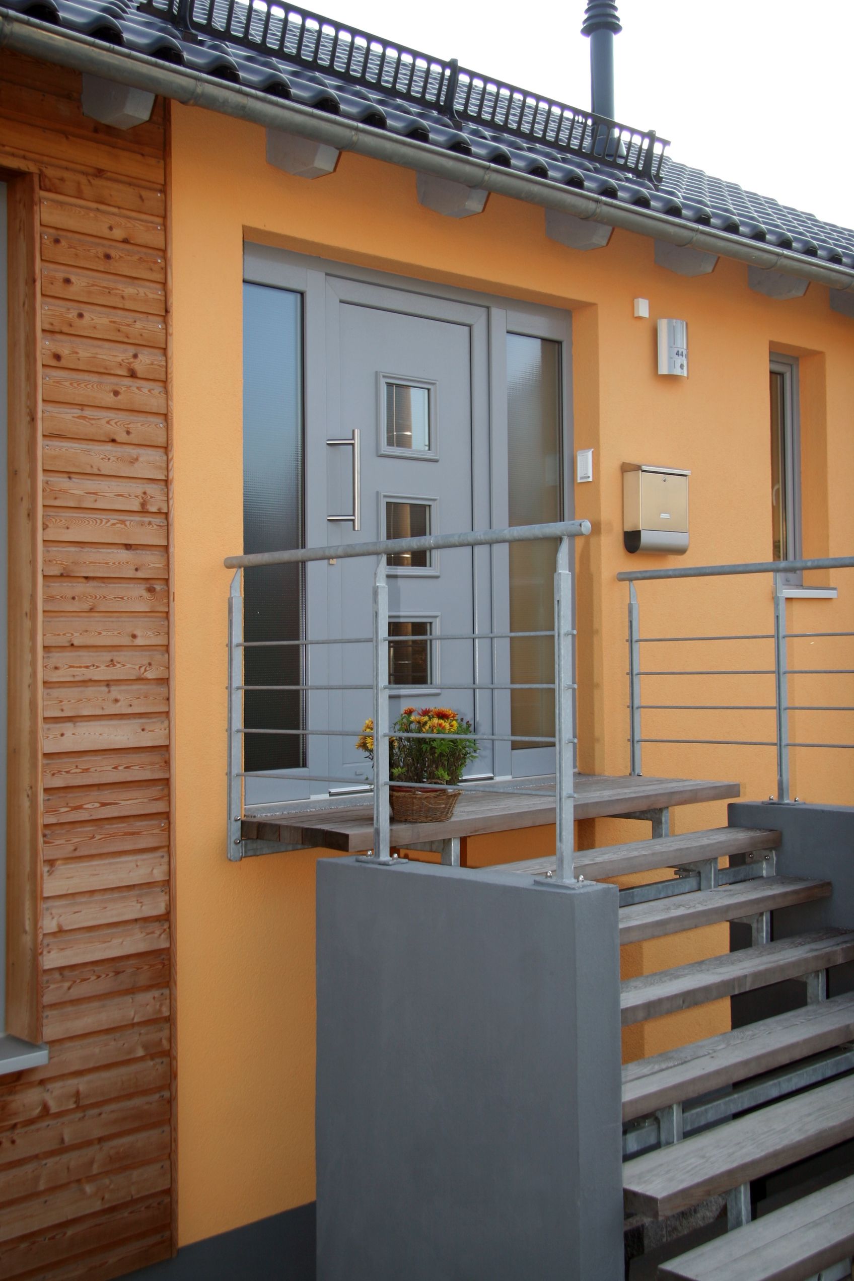 Orangefarbenes Gebäude mit grauer Tür und Treppe; Metallgeländer; Holzverkleidung und -treppe; Briefkasten.