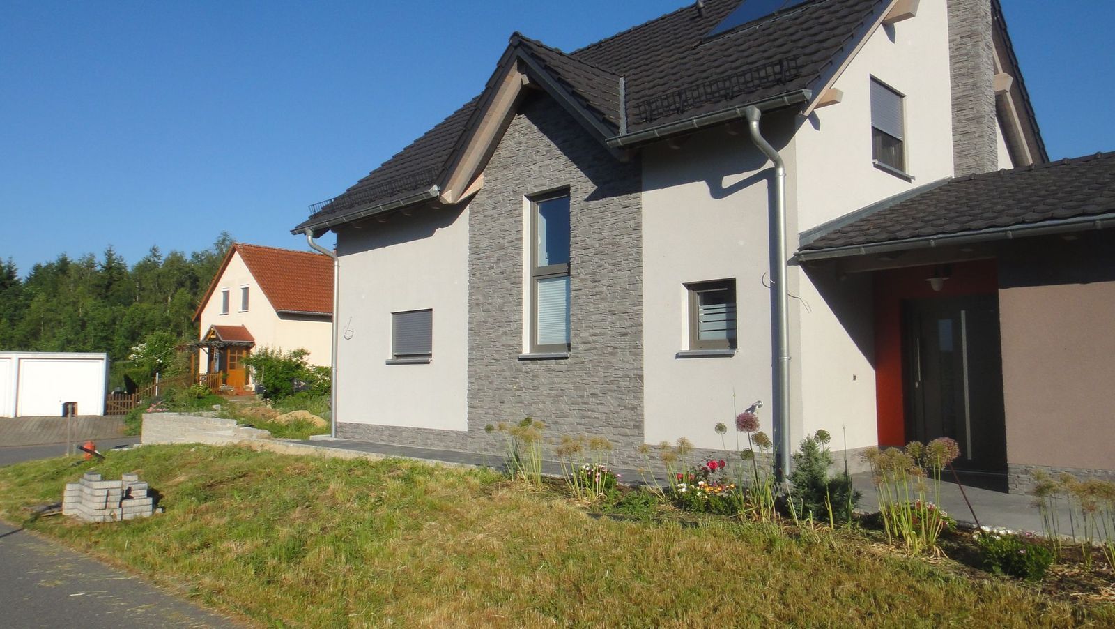 Modernes Haus mit Stein- und Stuckfassade, neben einem anderen Haus und einer Grünfläche, blauer Himmel.