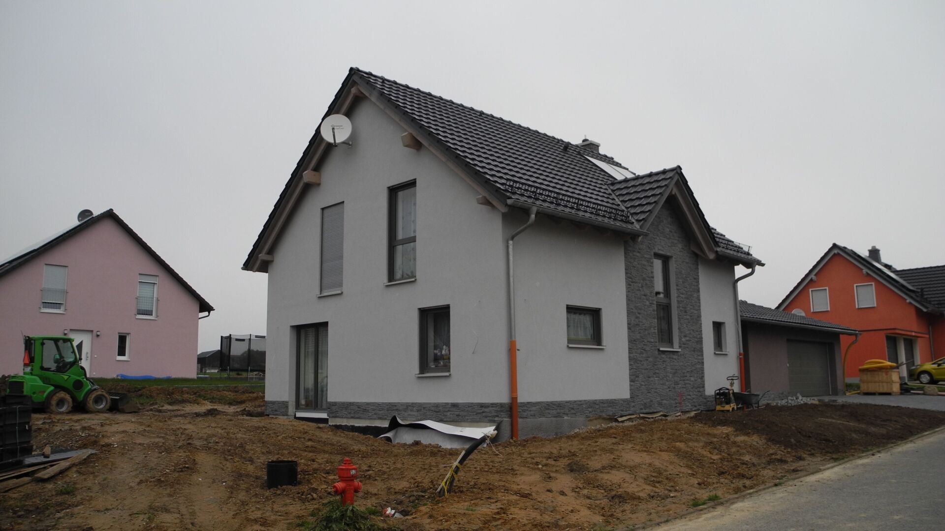 Zweistöckiges Haus mit grauer Fassade und schwarzem Dach. Im Bau, zwei weitere Häuser in der Nähe. Bewölkter Himmel.