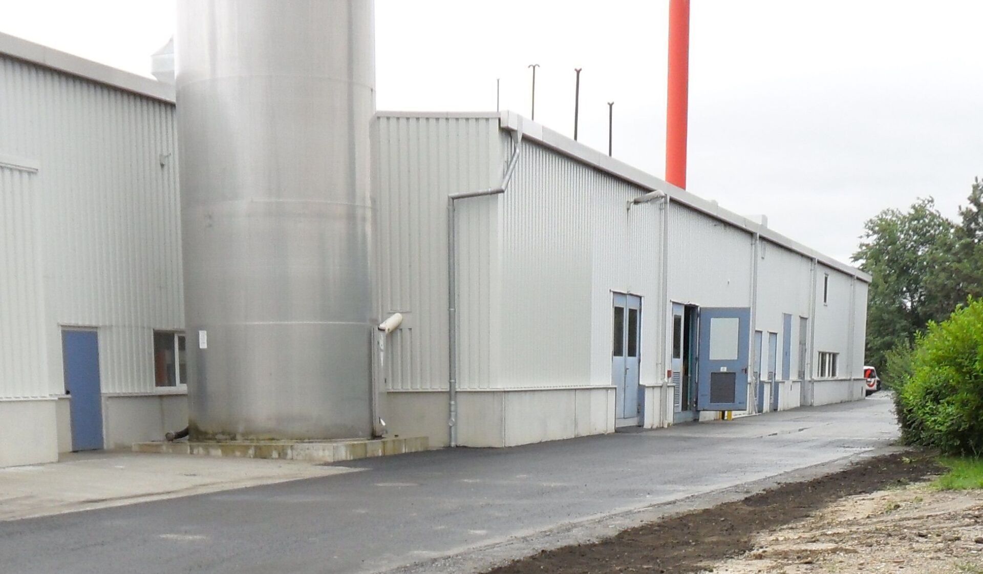 Industriegebäude mit einem großen silbernen Zylinder, einem roten Schornstein und einer dunklen Asphaltstraße.
