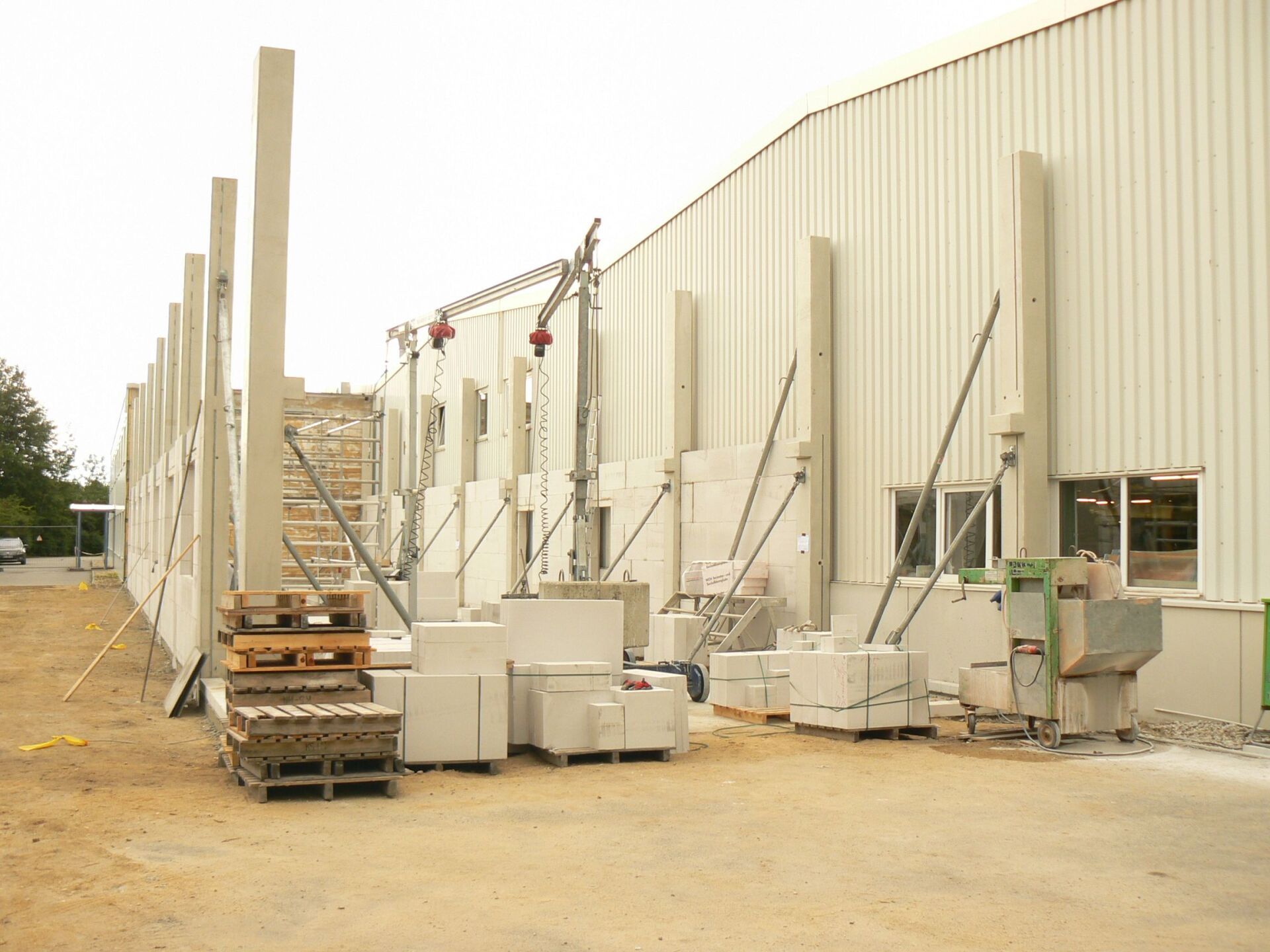 Baustelle mit Betonpfeilern und Gebäudefassade. Paletten, Blöcke und Kräne sind vorhanden.