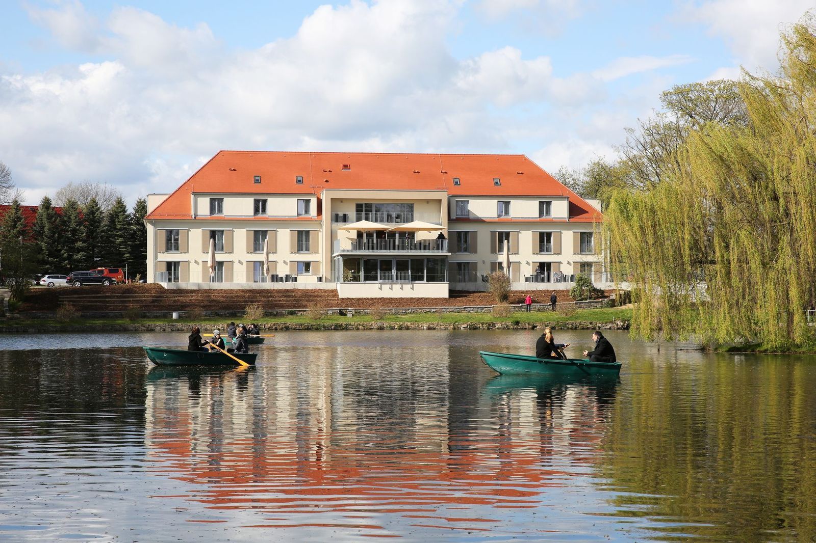 Großes Gebäude mit orangefarbenem Dach, das sich im See spiegelt; Menschen in Ruderbooten.