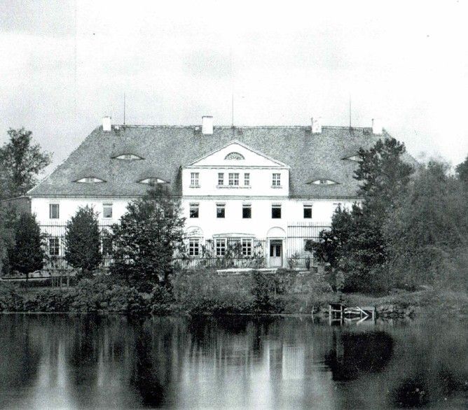 Großes weißes Herrenhaus mit rotem Ziegeldach, das sich in einem See spiegelt, eingebettet in ein Waldgebiet.