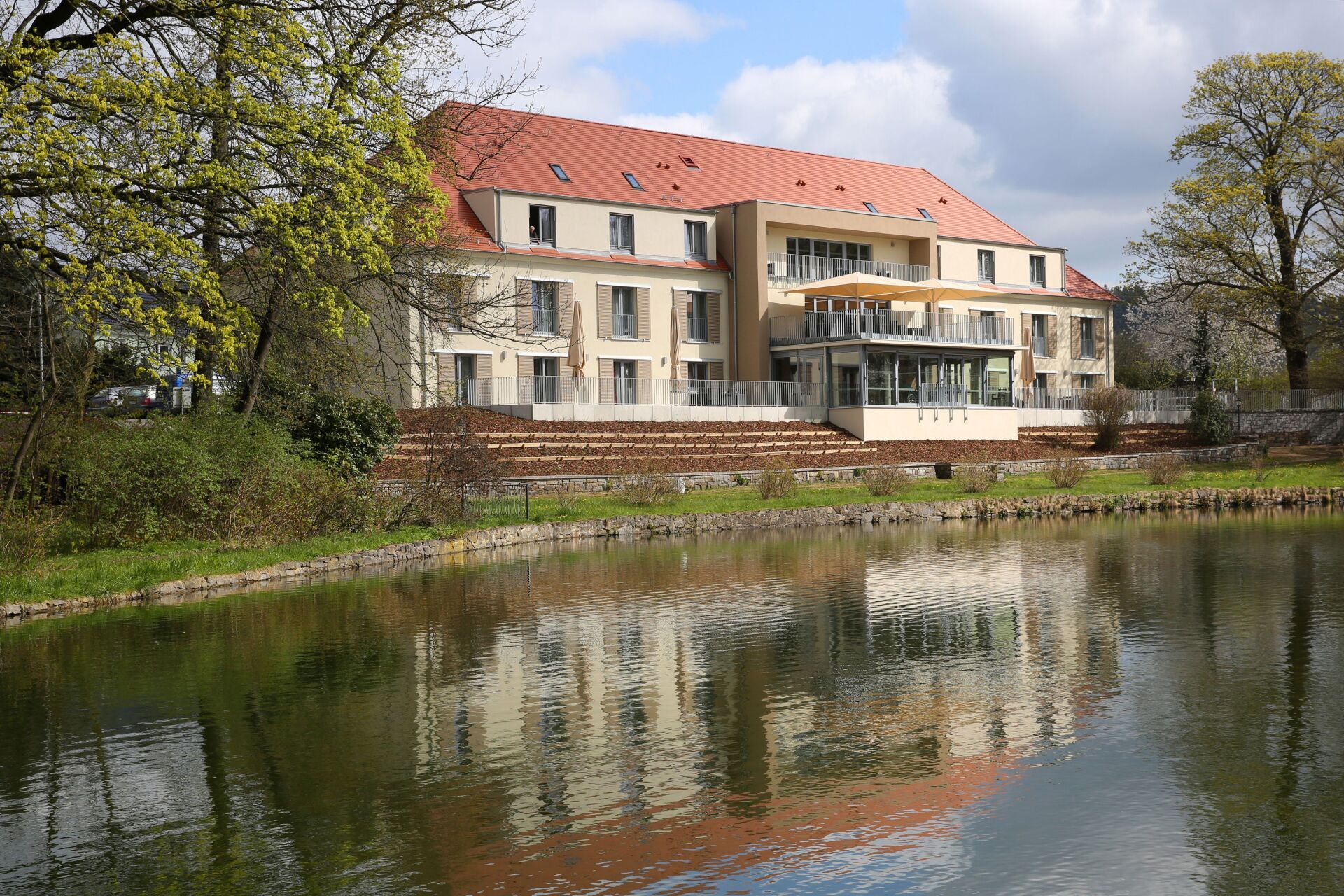 Großes Gebäude mit rotem Dach, beiger Fassade und verglastem Bereich, das sich im ruhigen Wasser spiegelt.