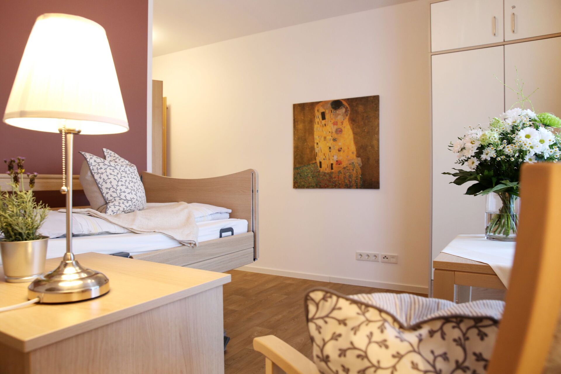 Gemütliches Schlafzimmer mit Bett, Kunstwerken, Lampe und Blumen; das Zimmer hat eine warme Farbpalette.