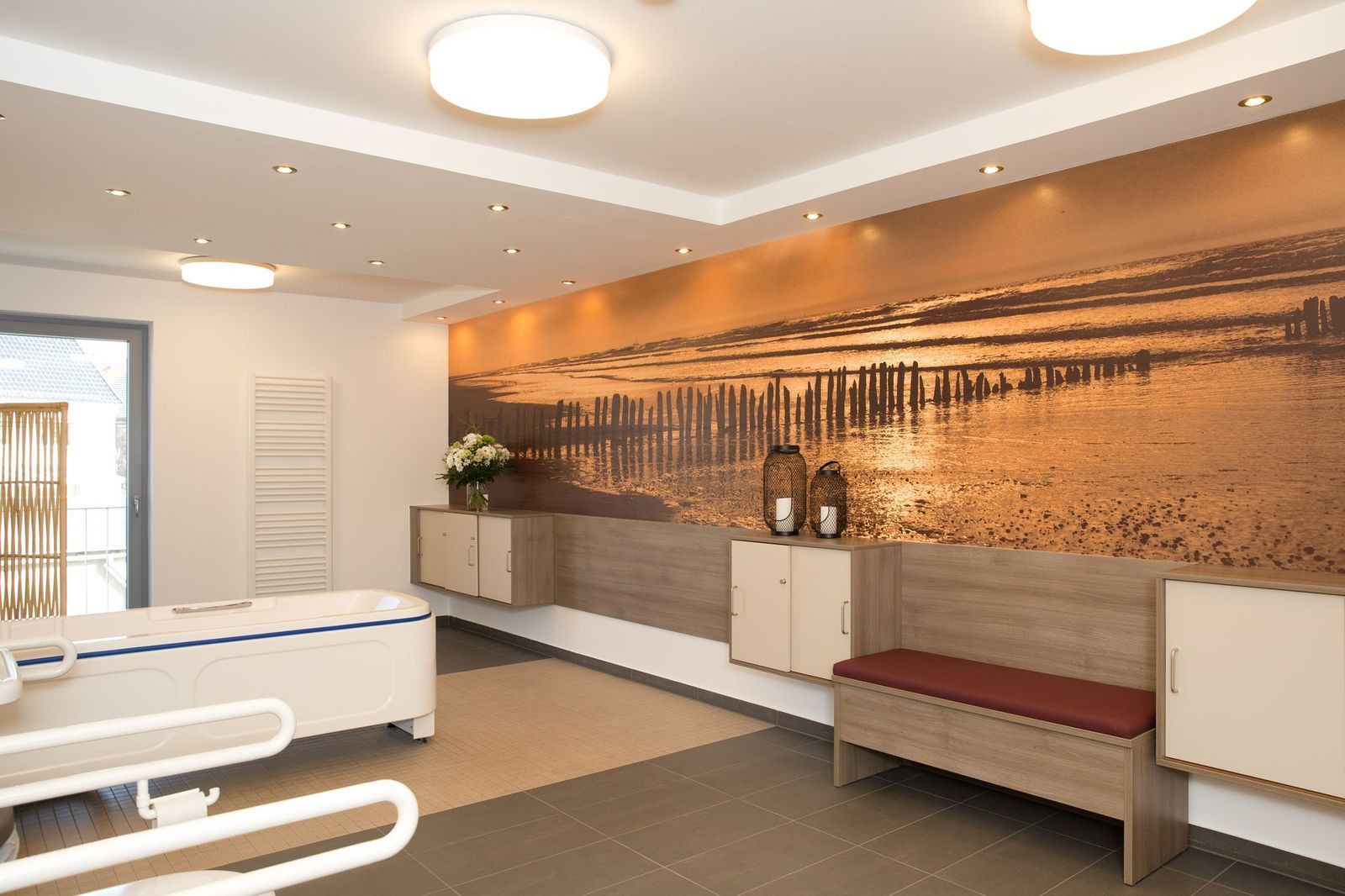 Ein Wellnessraum mit einem Wandgemälde einer Strandszene, Sitzgelegenheiten und einer therapeutischen Badewanne mit Haltegriffen.