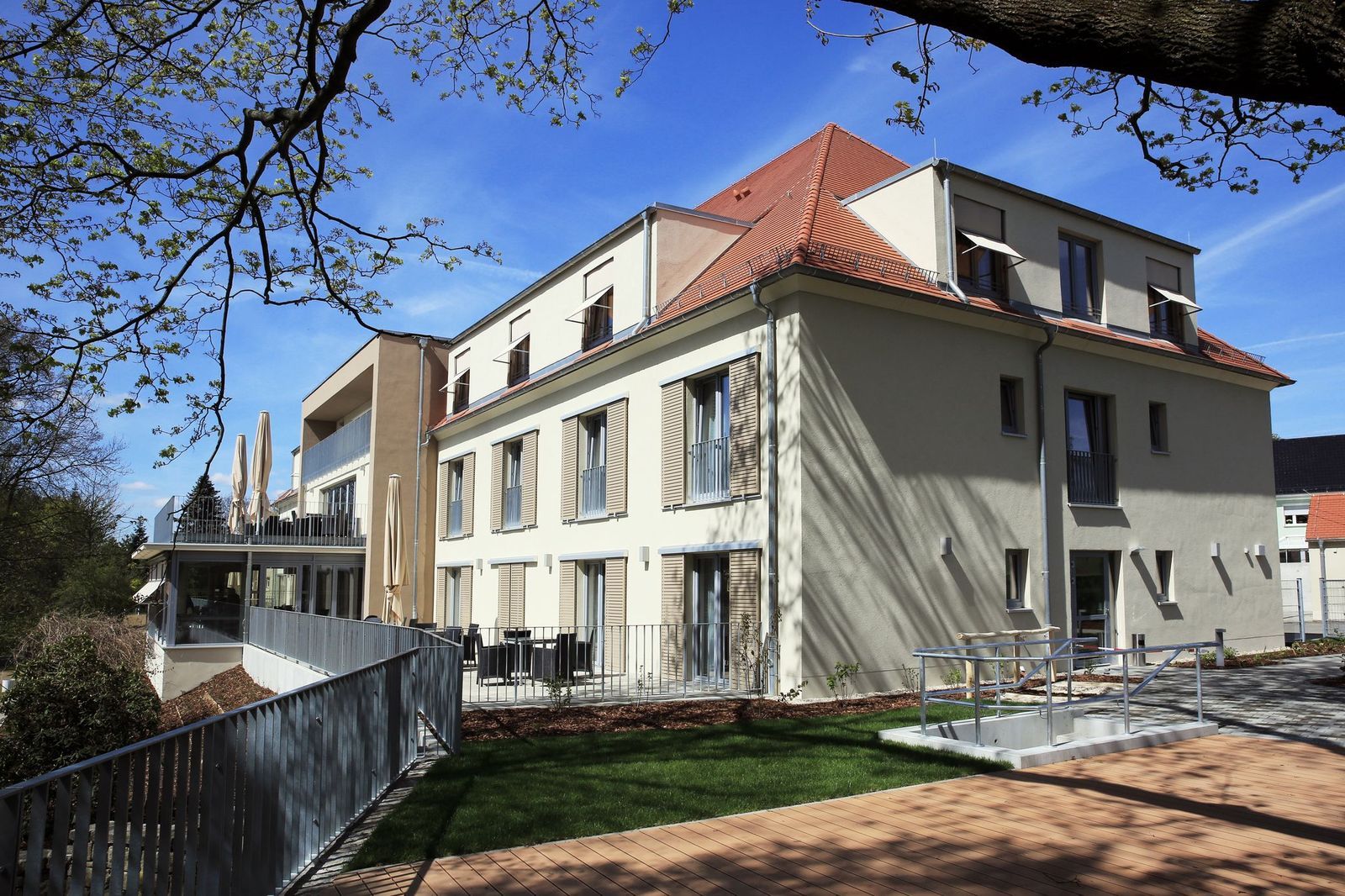 Dreistöckiges Gebäude mit beiger Fassade, rotem Ziegeldach und Außensitzplätzen an einem sonnigen Tag.