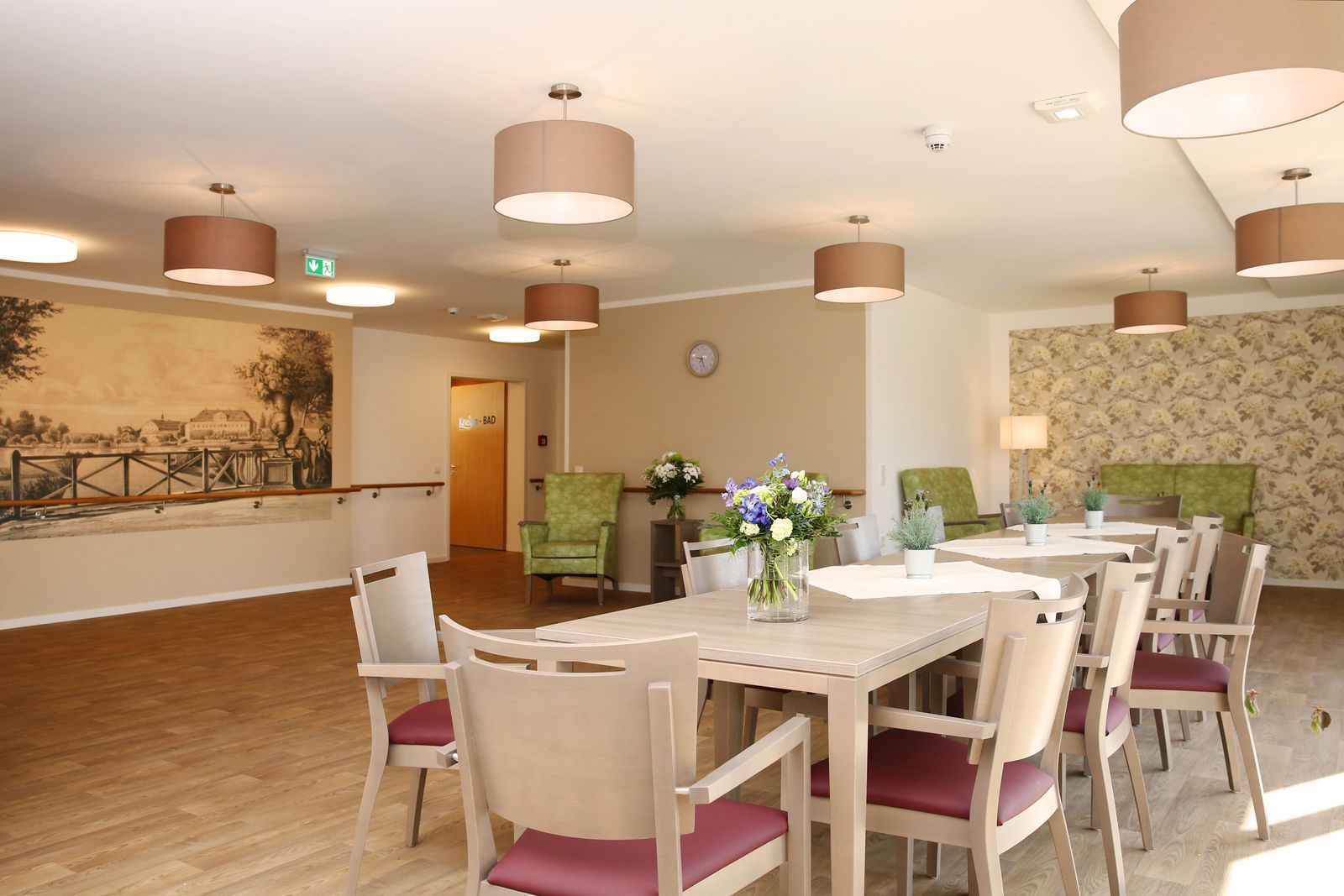 Esszimmer mit Tischen, Stühlen und Pendelleuchten. Ein Wandgemälde und eine gemusterte Tapete sind zu sehen.