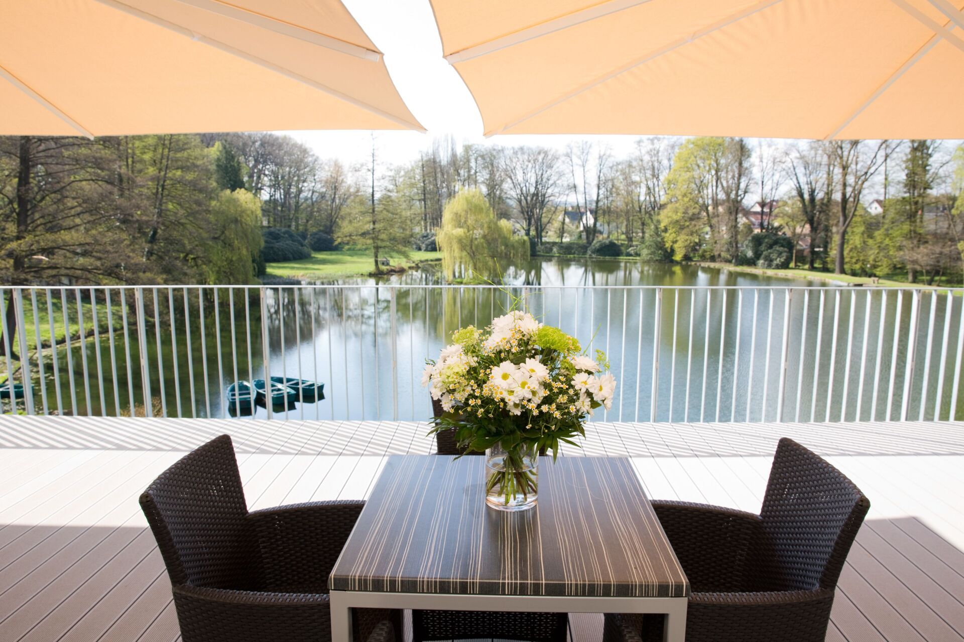 Terrasse mit Blick auf einen See, mit Tisch, Stühlen und Blumen. Cremefarbene Markise.