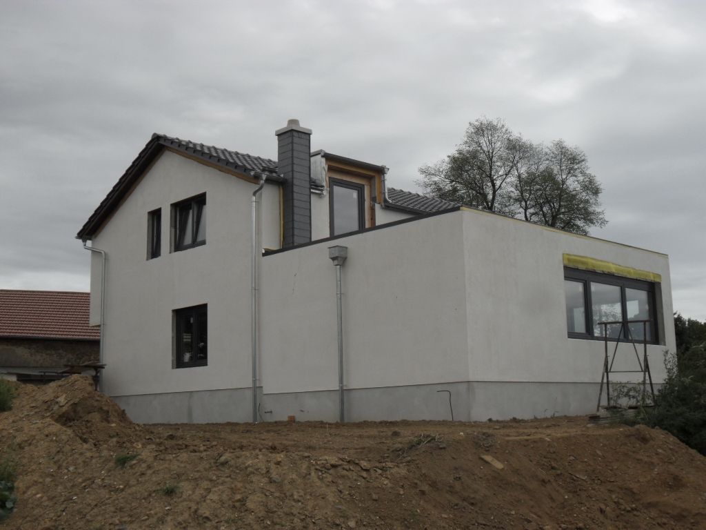 Weißes Haus im Bau, mit Schornstein und grauem Dach, vor bewölktem Himmel.
