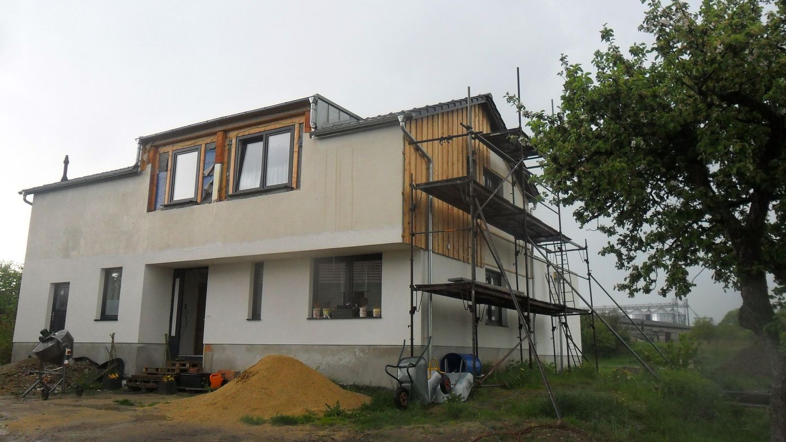 Zweistöckiges Haus im Bau. Gerüst, Holzverkleidung und weißer Putz. Baum im Hintergrund.