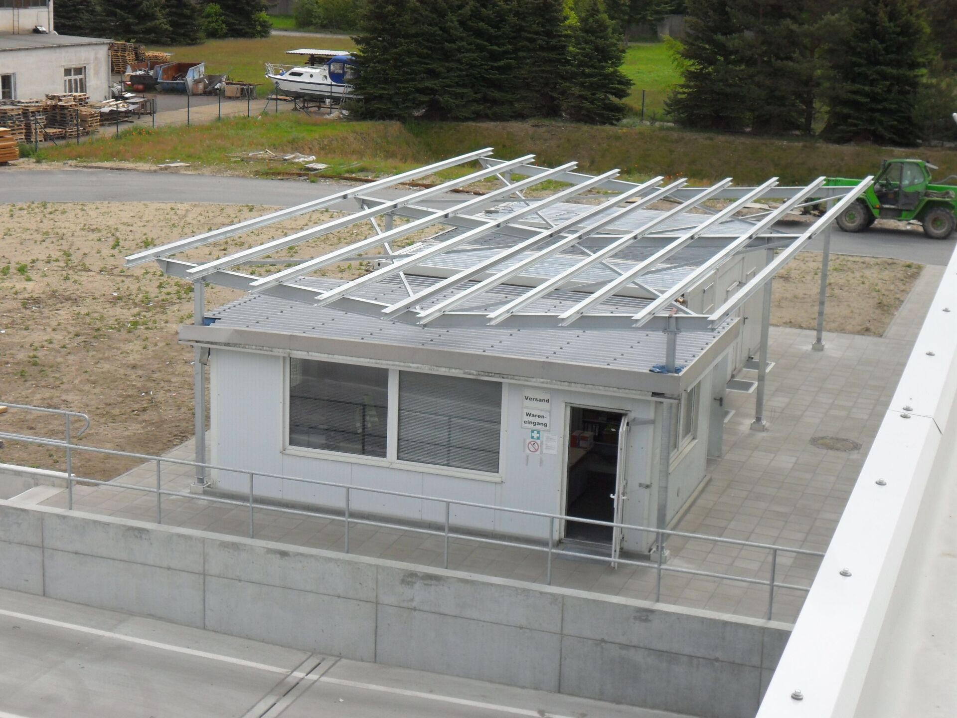 Kleines Gebäude mit Solarpaneelen auf dem Dach, von oben betrachtet, umgeben von Beton und Gras.