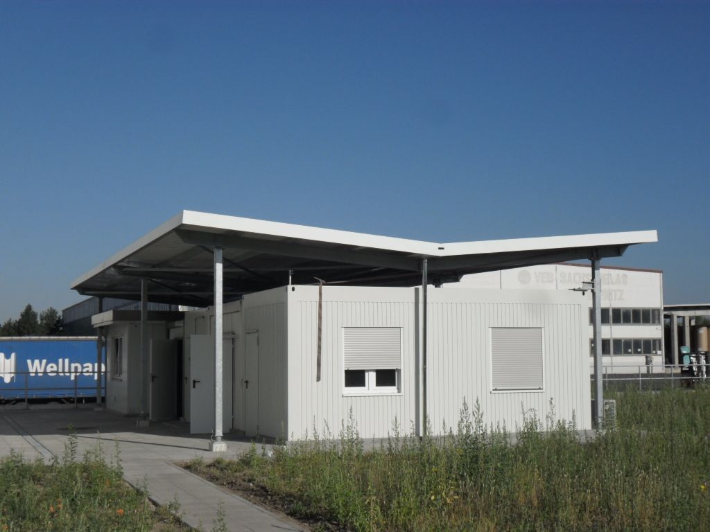 Weißes Industriegebäude mit schrägem Dach, zwei Fenster mit Jalousien, unter blauem Himmel.