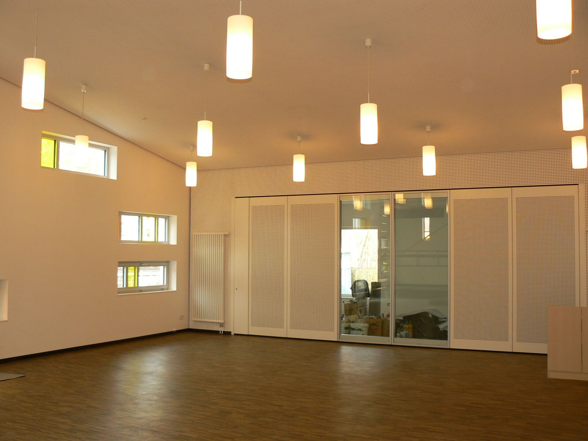 Helles, leeres Zimmer mit zylinderförmigen Hängelampen. Ausgestattet mit Fenstern, Schiebetüren und weißen Wänden mit gemusterten Paneelen.