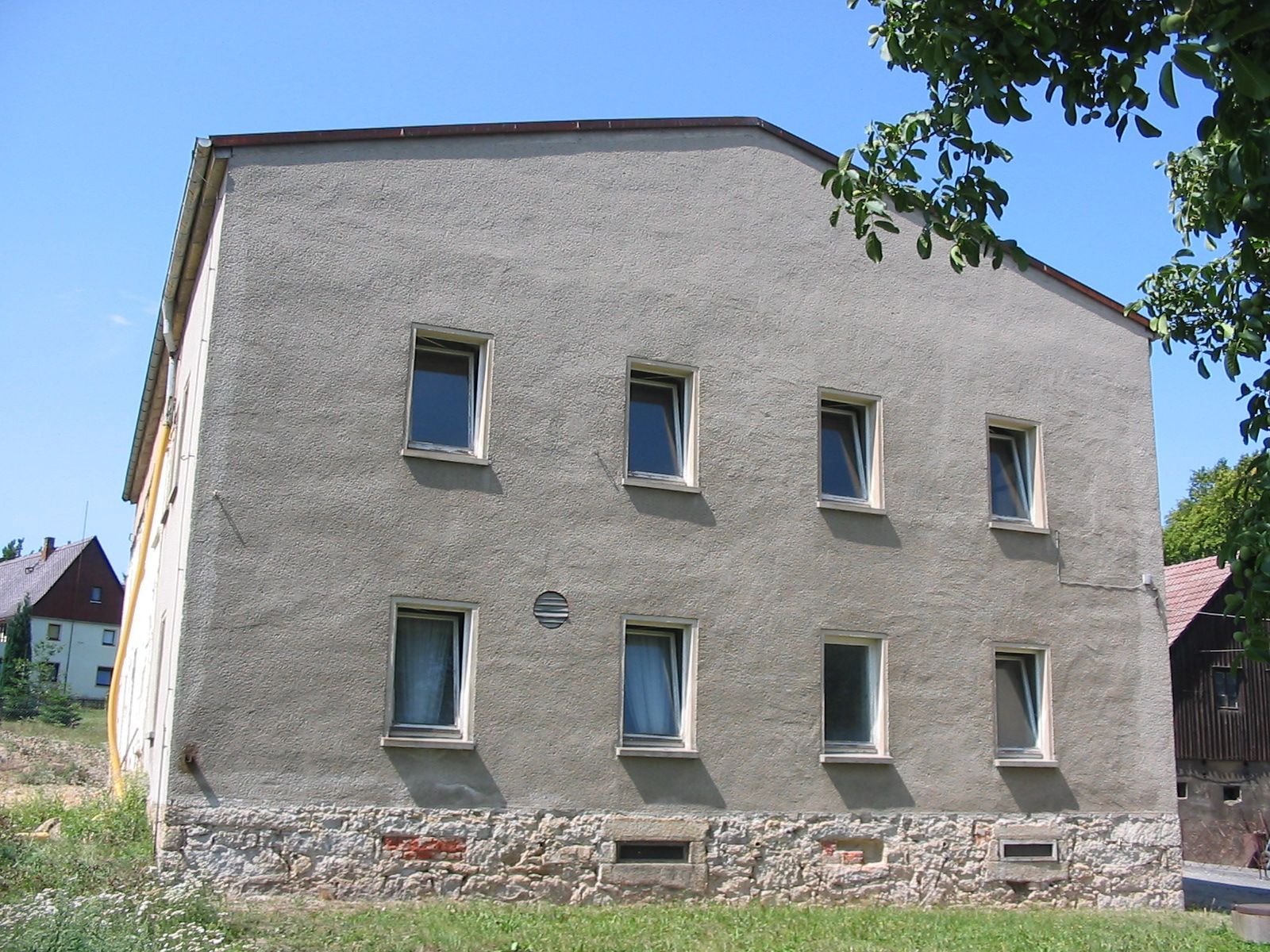 Zweistöckiges graues Gebäude mit Steinfundament und acht Fenstern. Grünes Gras im Vordergrund, blauer Himmel.