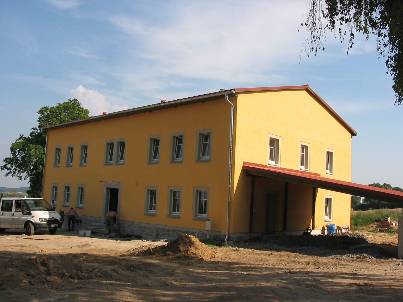 Gelbes zweistöckiges Gebäude mit roter Markise. Ein weißer Lieferwagen und Menschen stehen an einem sonnigen Tag draußen.
