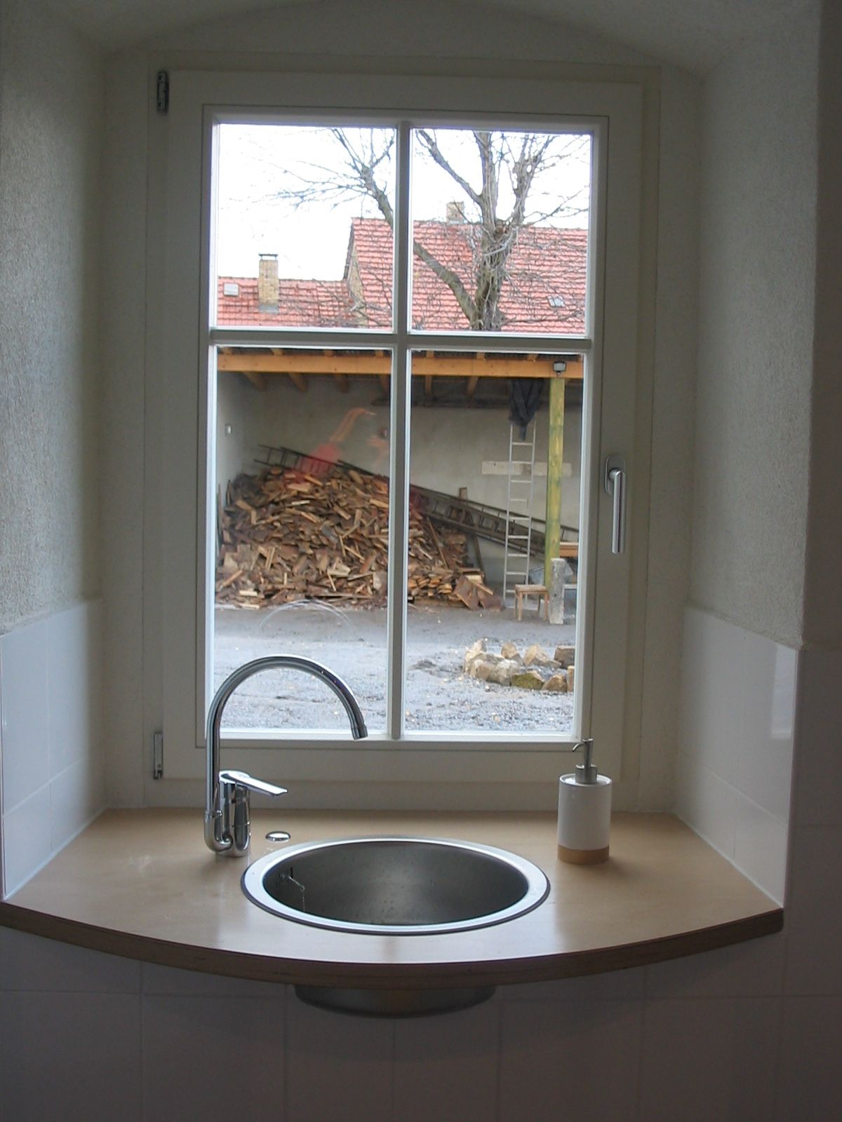 Waschbecken und Wasserhahn vor einem Fenster mit Blick auf einen Hof mit gestapeltem Brennholz.