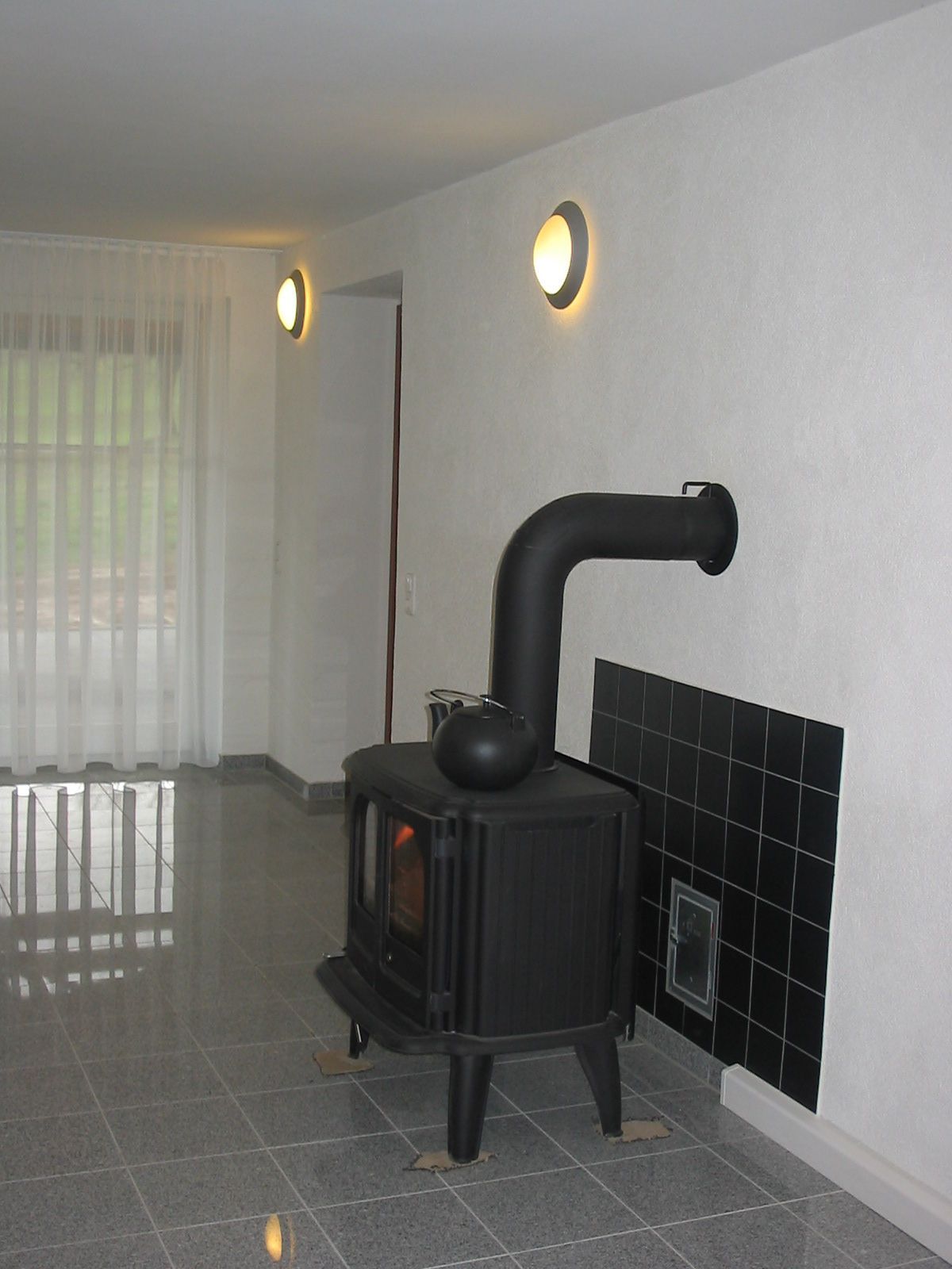 Schwarzer Holzofen mit gebogenem Schornstein vor weißer Wand mit gefliester Rückwand, zwei Wandleuchten und verhängtem Fenster.