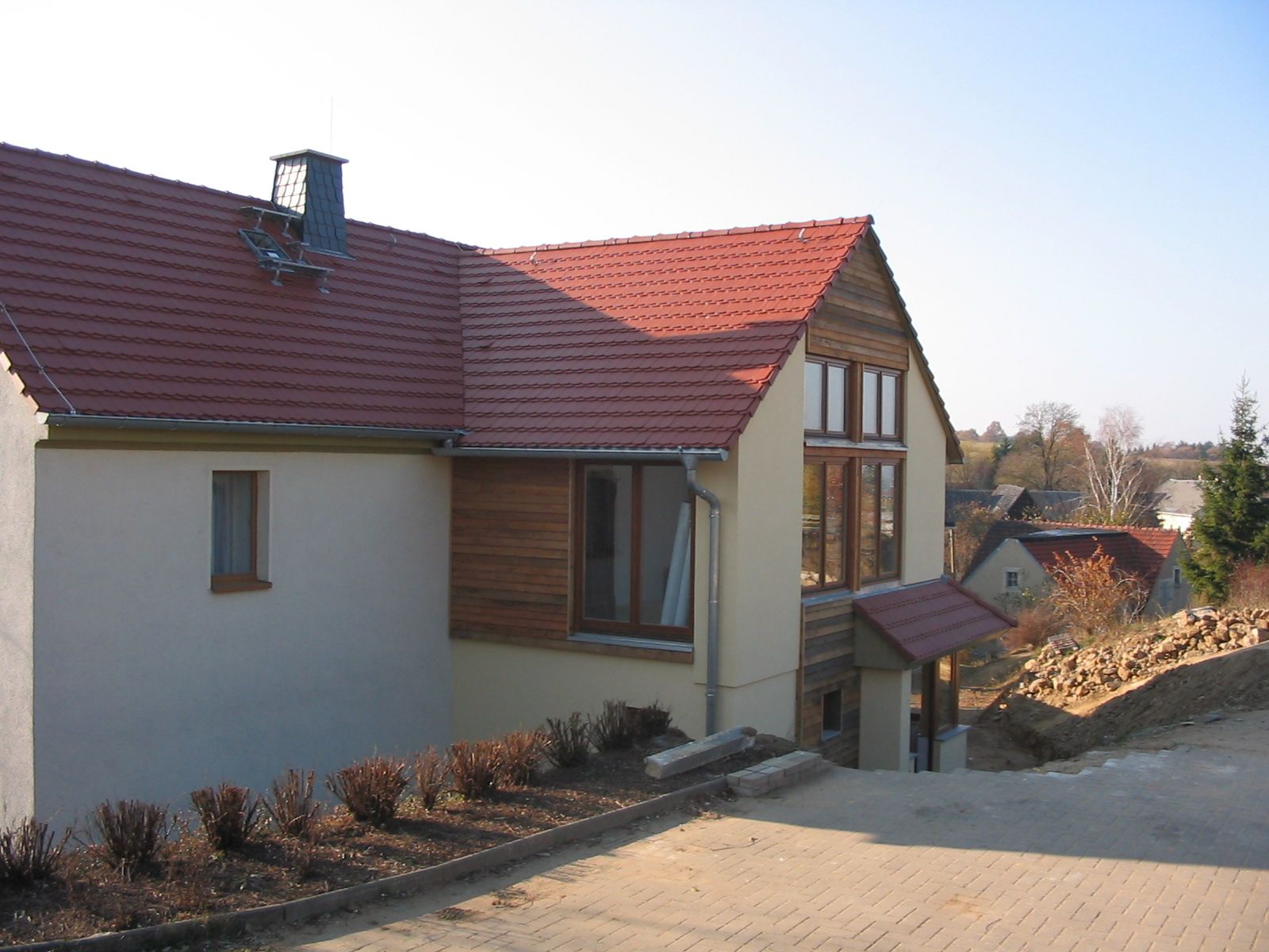 Beiges Haus mit rotem Ziegeldach, Holzelementen und großen Fenstern an einer abfallenden Straße.