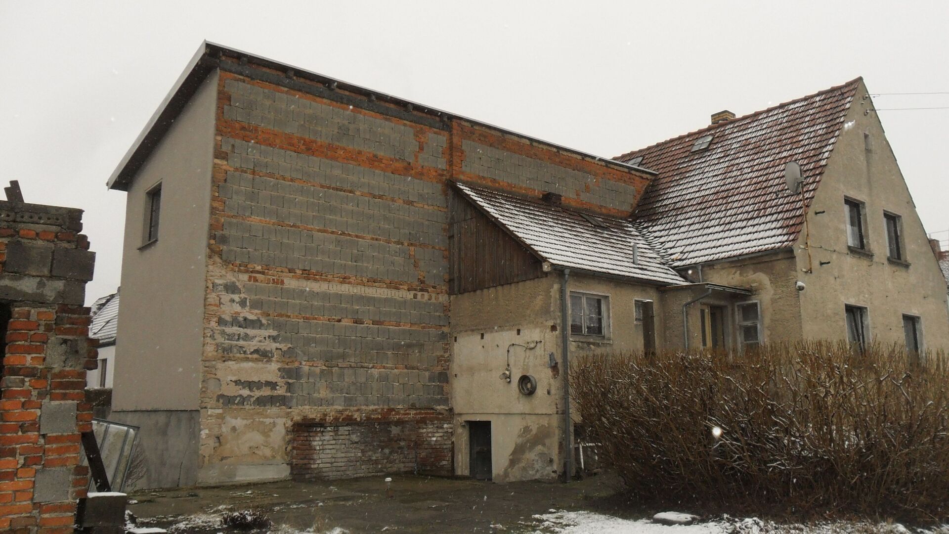 Teilweise renovierte Gebäudefassade aus Ziegeln, Beton und rotem Ziegeldach, bedeckt mit Schnee.