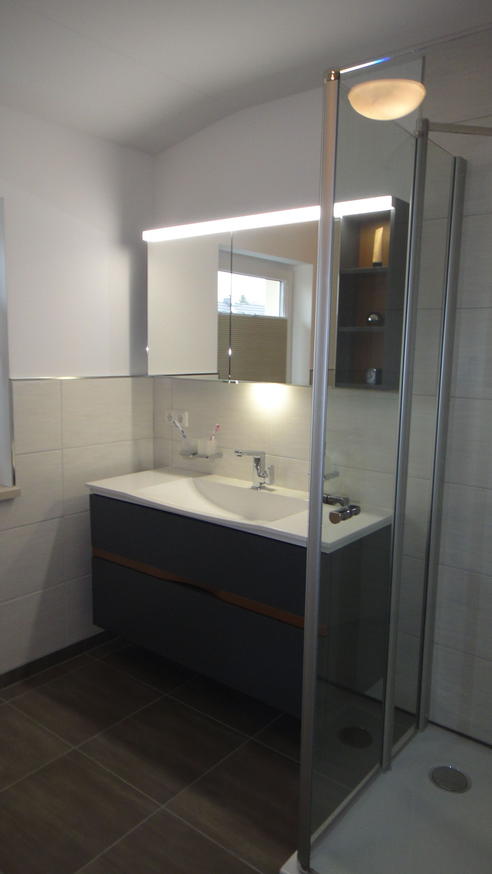 Badezimmer mit dunkelgrauem Waschtisch, Waschbecken und Spiegel mit Deckenbeleuchtung; Duschkabine aus Glas.
